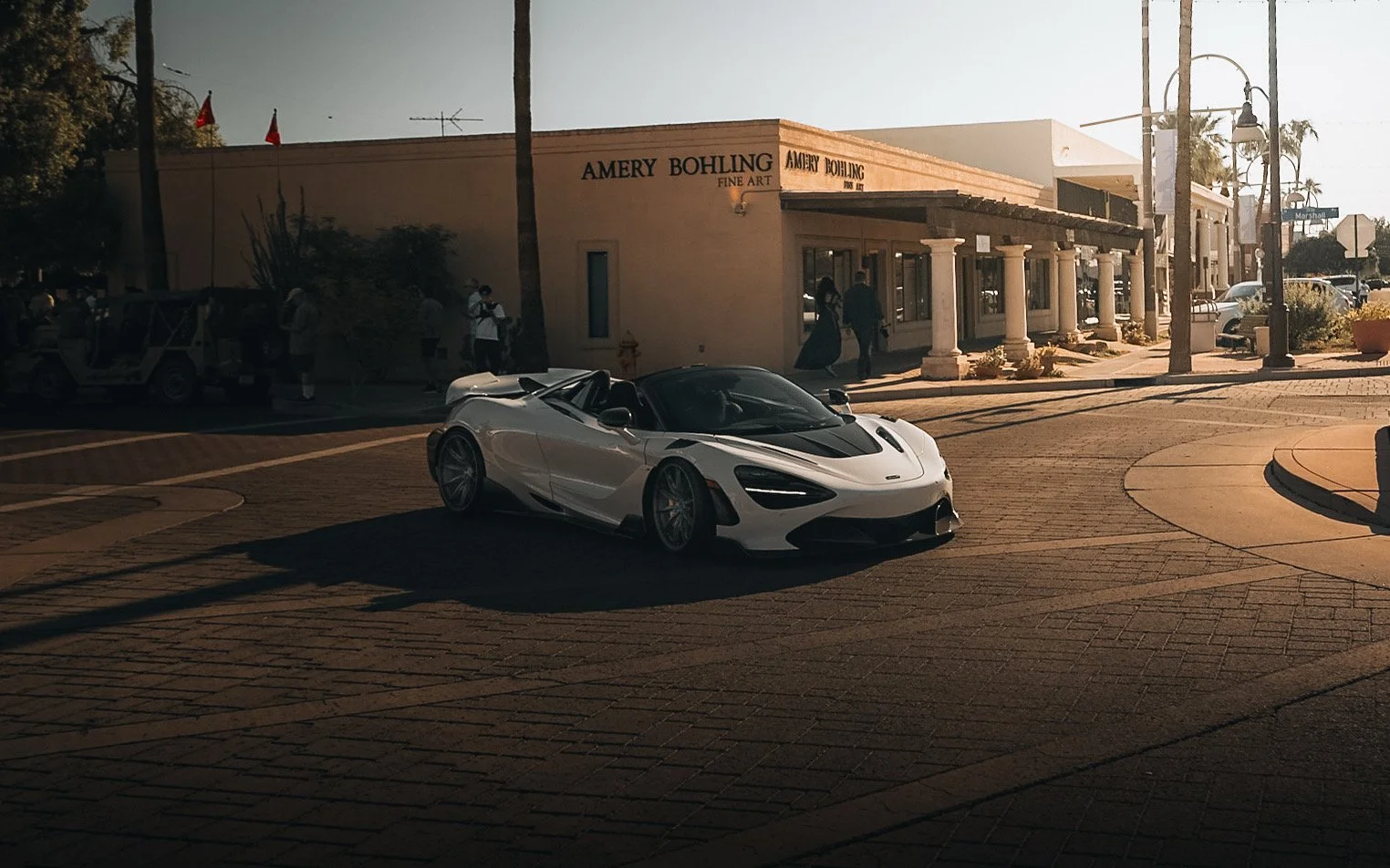 A white luxury sports car parked on a city street in front of a fine art gallery. The gallery has a sign that reads "Amery Bohling Fine Art". The scene is during daylight with people walking and a few vintage-style street lamps.
