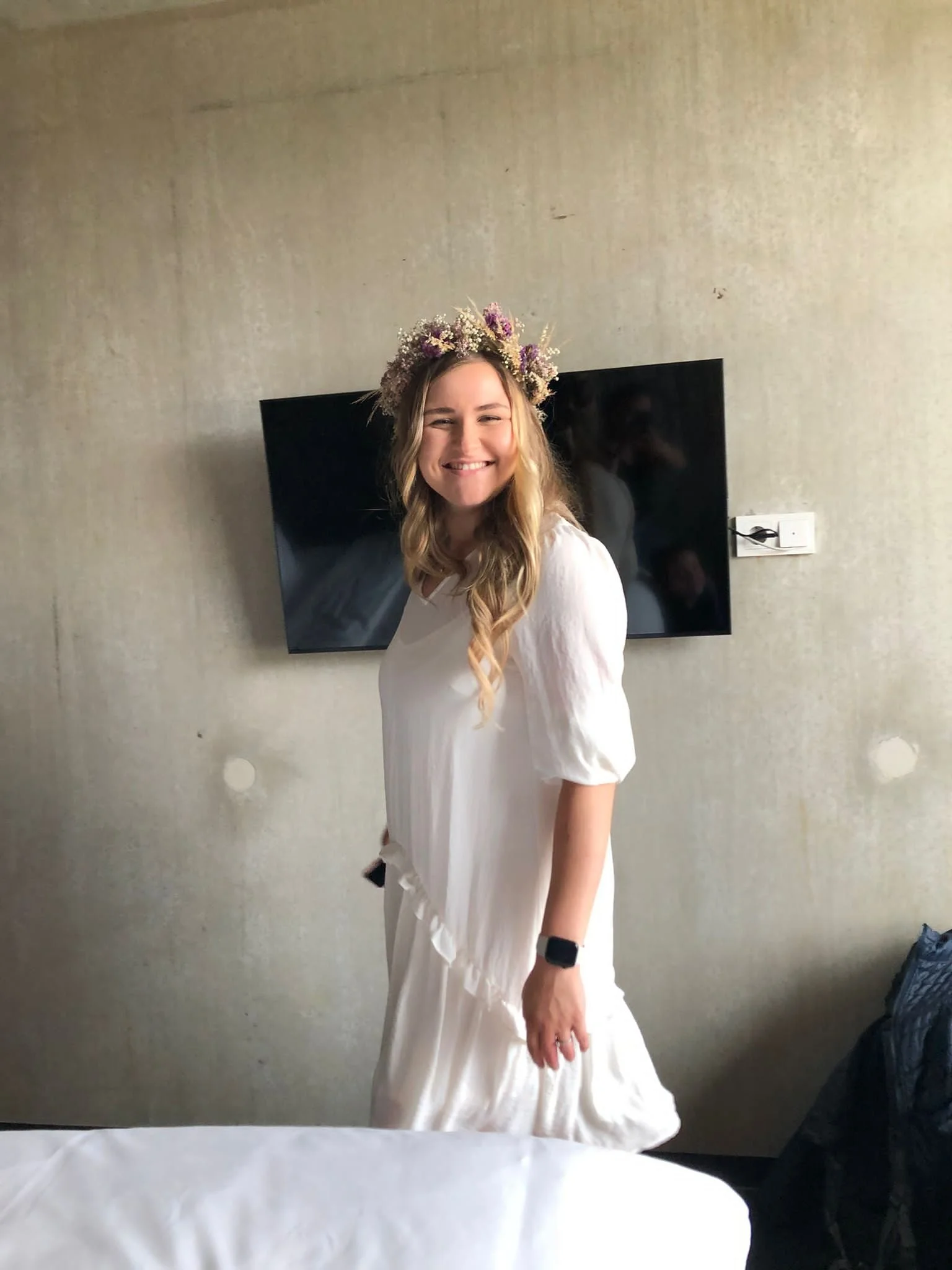 Junge Frau mit Blumenkranz auf dem Kopf, in weißem Kleid, leicht lächelnd, im Hintergrund ein Fernseher an einer grauen Wand.