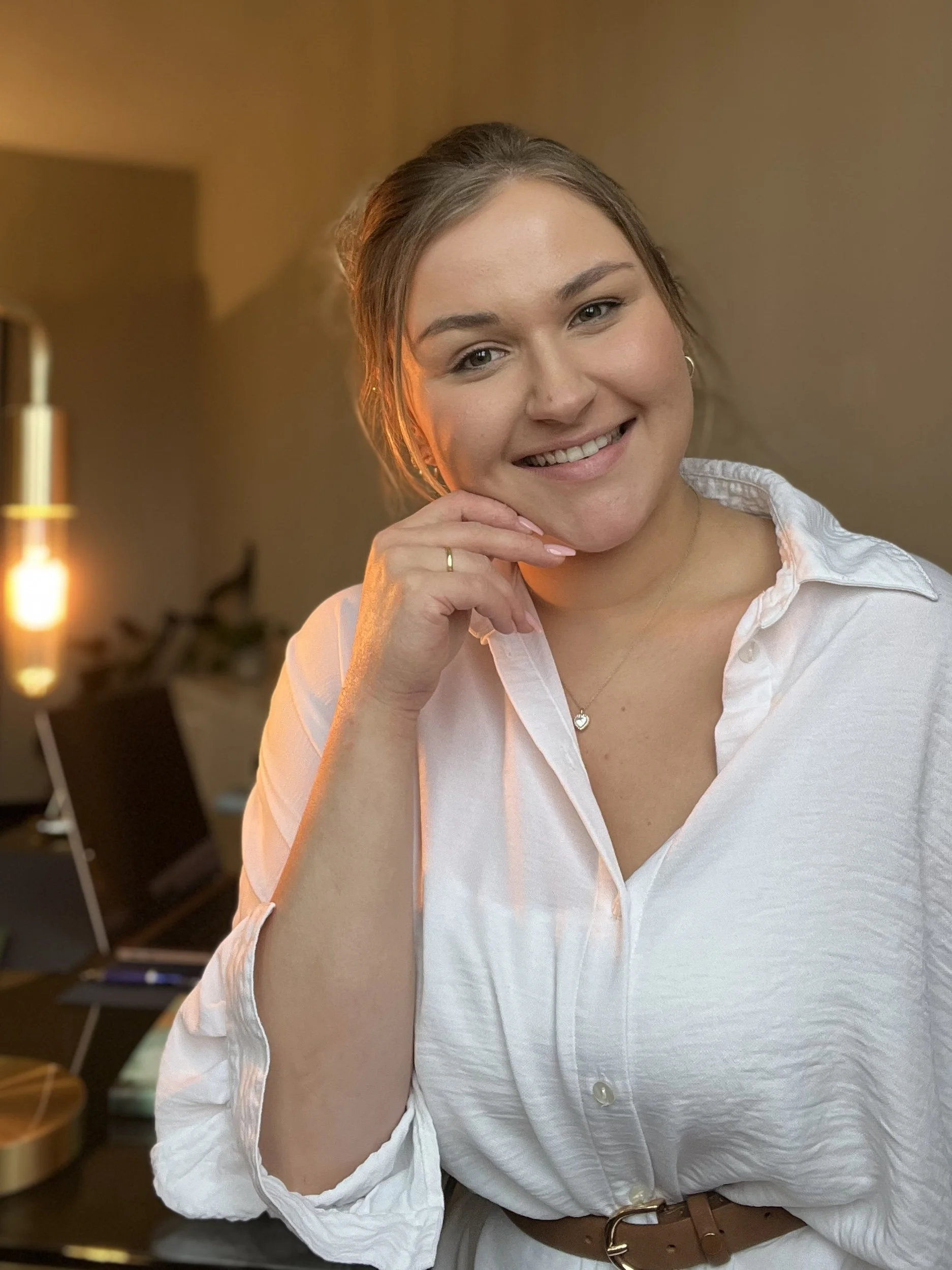 Lächelnde junge Frau mit braunen Haaren, weiße Bluse, im Hintergrund ein Laptop auf einem Tisch, warmes Licht im Raum.