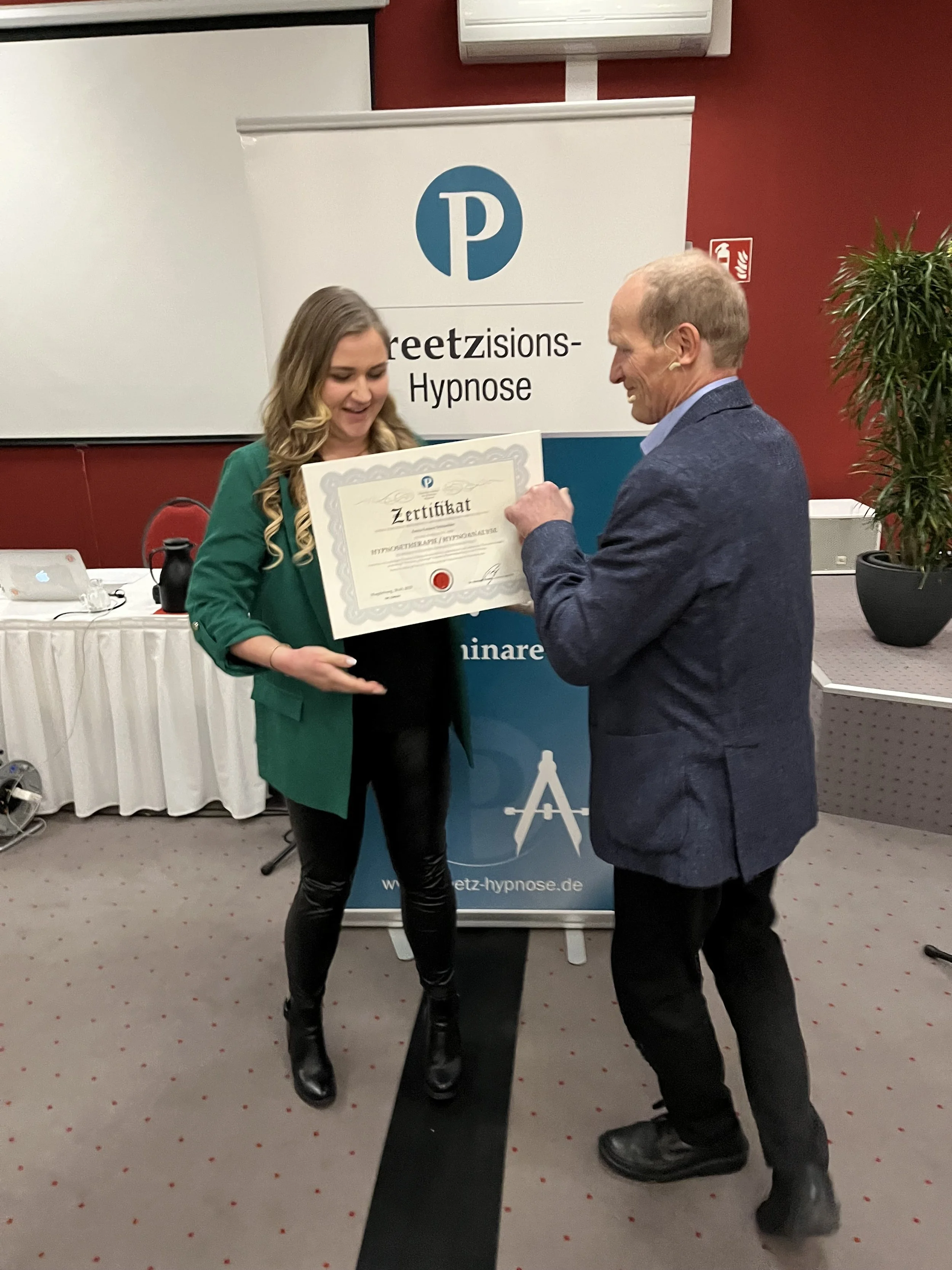 Ein Mann überreicht einer lächelnden jungen Frau ein Zertifikat bei einer Veranstaltung. Im Hintergrund ein Banner mit dem Logo und dem Text 'Greetzisations-Hypnose'.