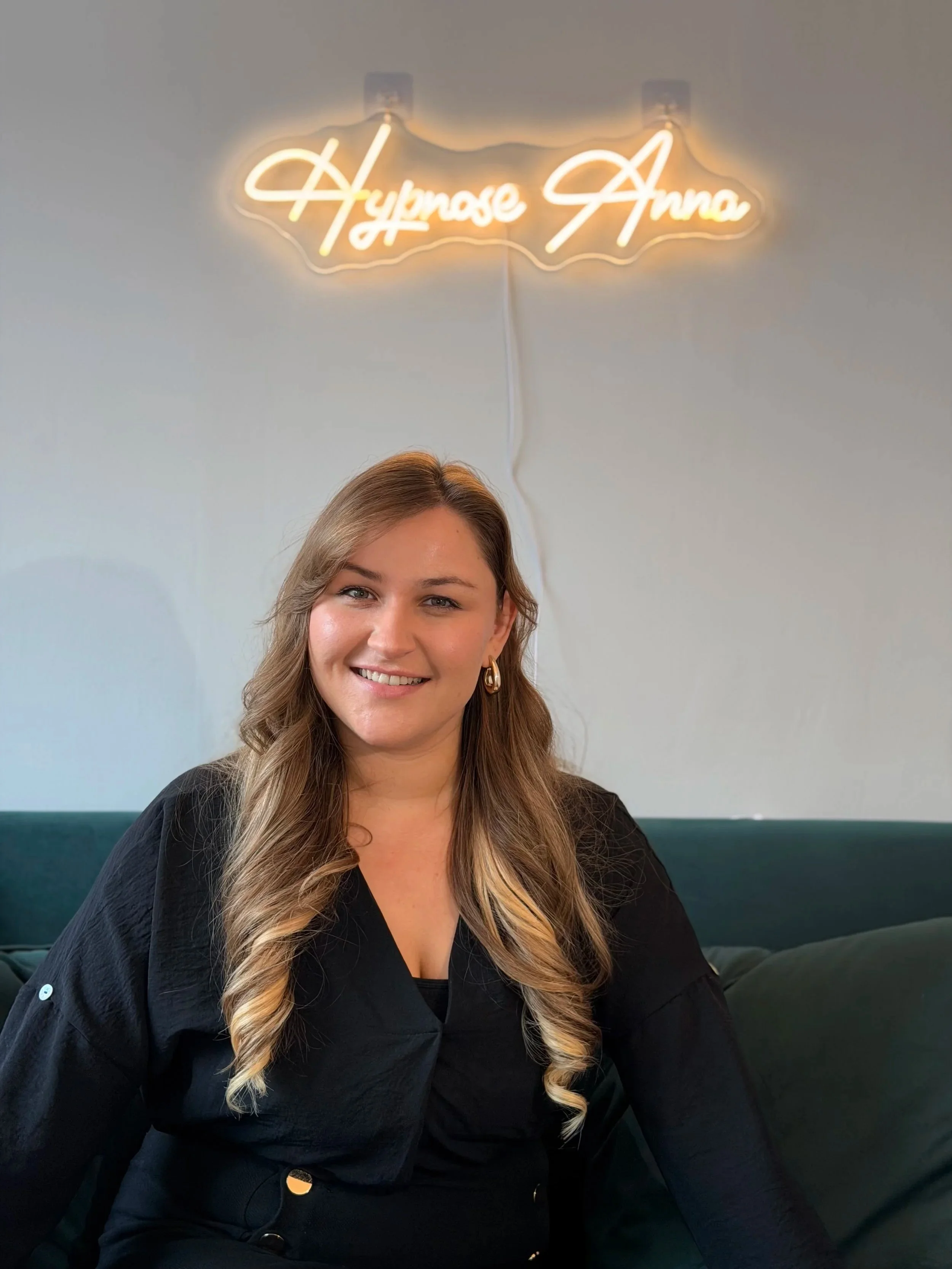 Eine Frau sitzt auf einer dunklen Couch vor einer grauen Wand mit einer leuchtenden Neon-Reklame, die 'Hymose Anna' heißt.