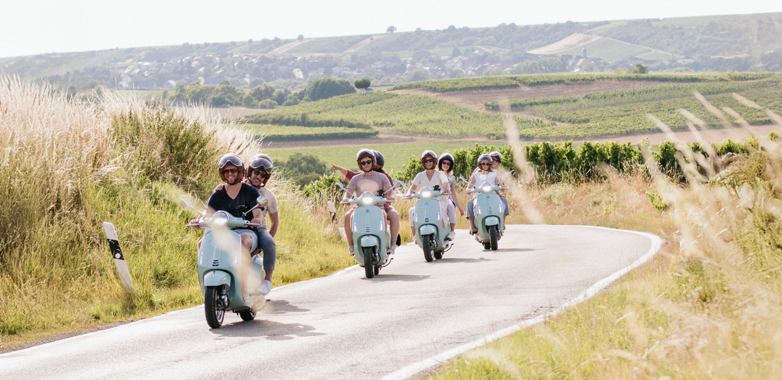 Gruppe auf mintfarbenen Vespas auf einer kurvigen Landstraße durch die Weinberge Rheinhessens