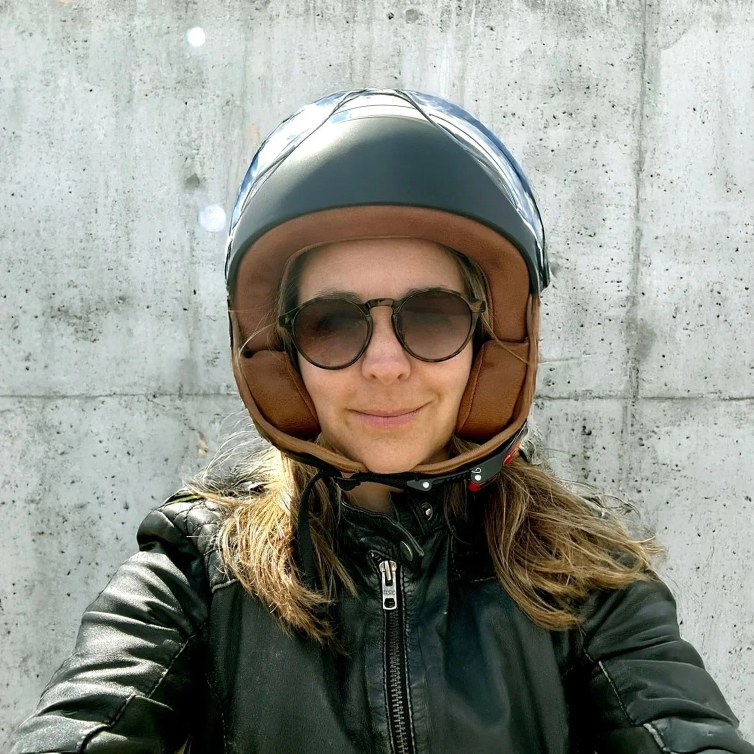 Junge Frau trägt Motorradhelm, Sonnenbrille und Lederjacke vor einer betonierten Wand.