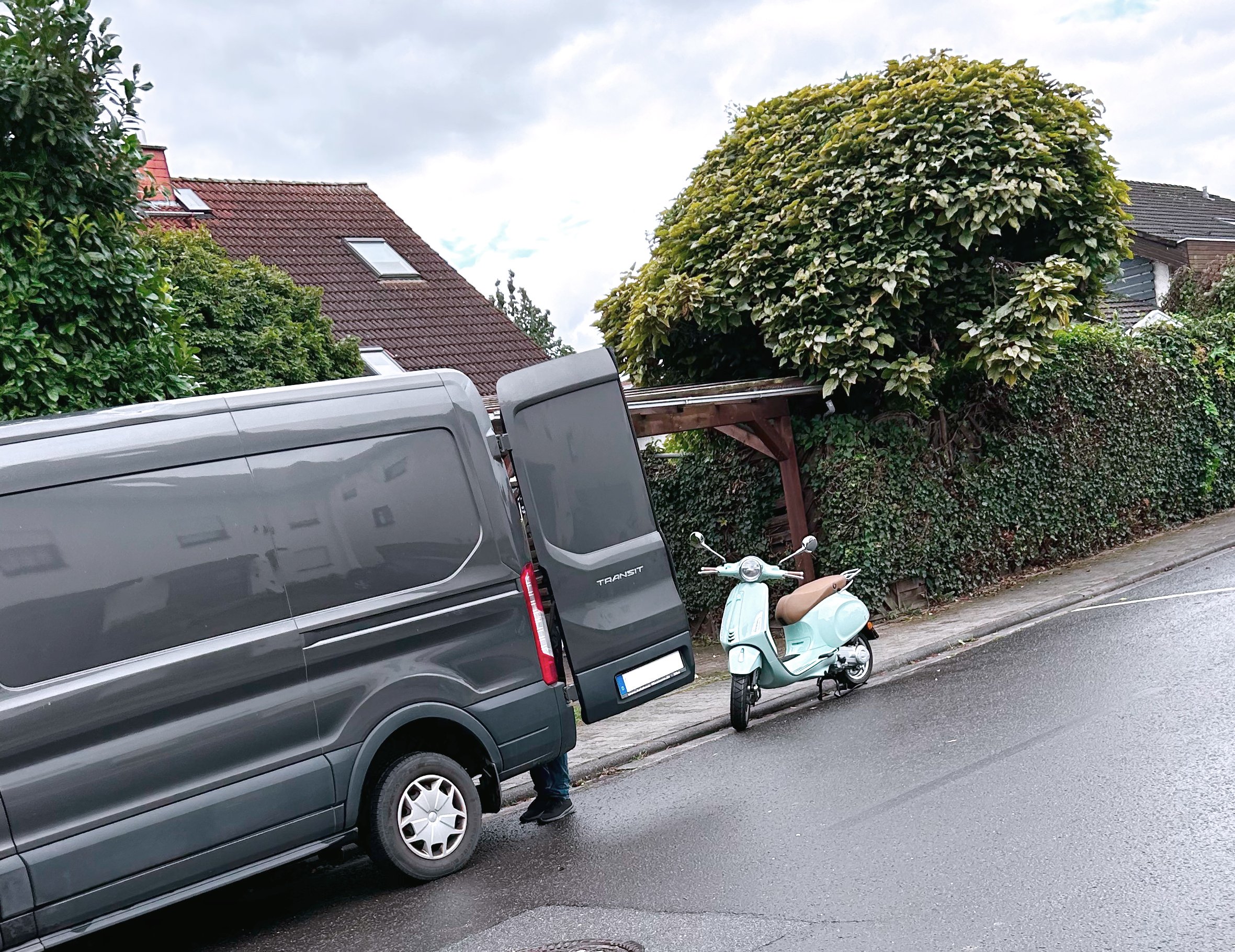 Ein grauer Lieferwagen parkt am Straßenrand, mit einer Person, die den Kofferraum öffnet, sichtbar. Ein weißes Motorroller steht neben dem Wagen. Im Hintergrund sind Häuser und ein großer Busch zu sehen.