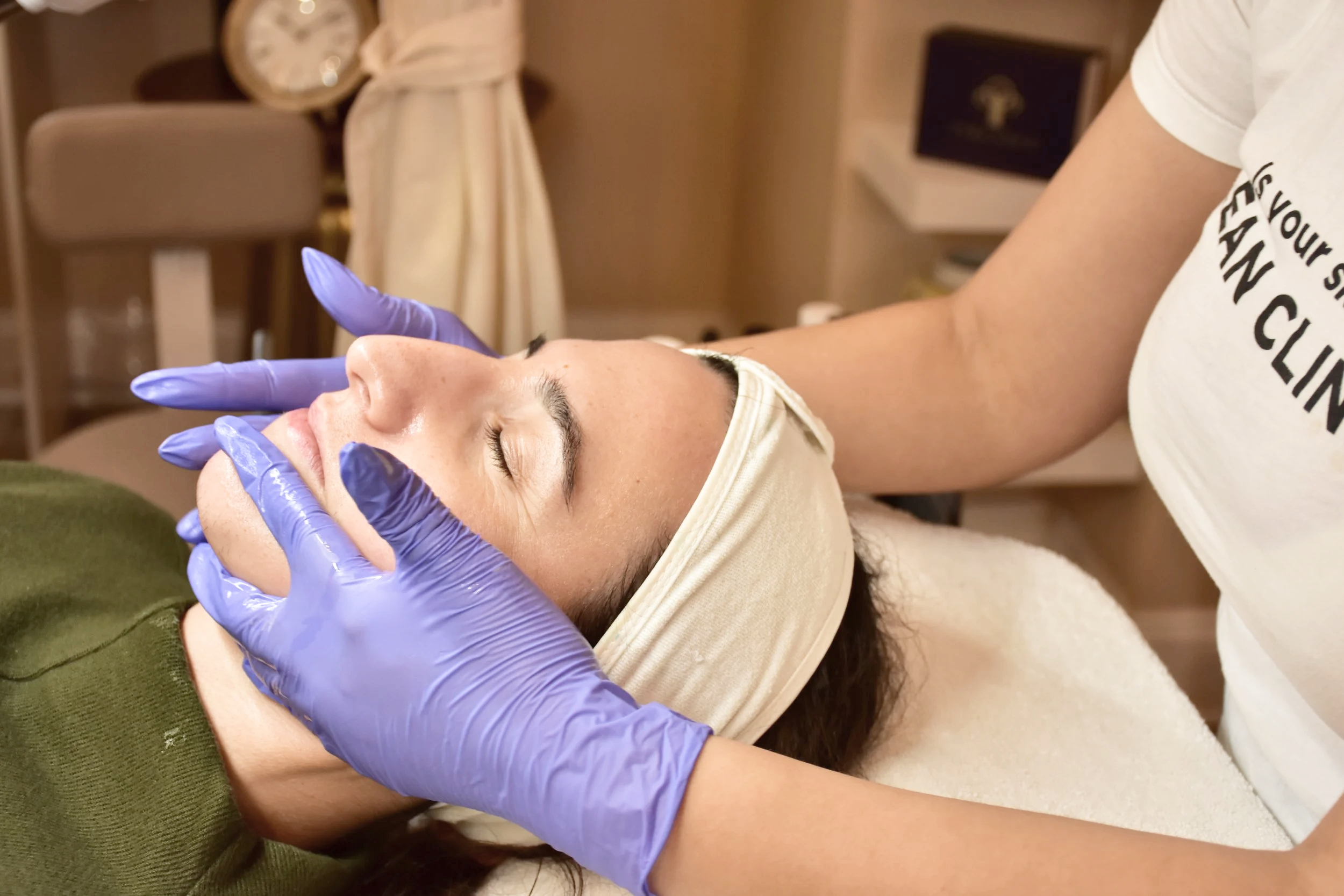 A woman receiving a facial treatment, lying down with her eyes closed, wearing a headband, while a skincare professional in purple gloves applies treatment to her face in a spa or clinic setting.