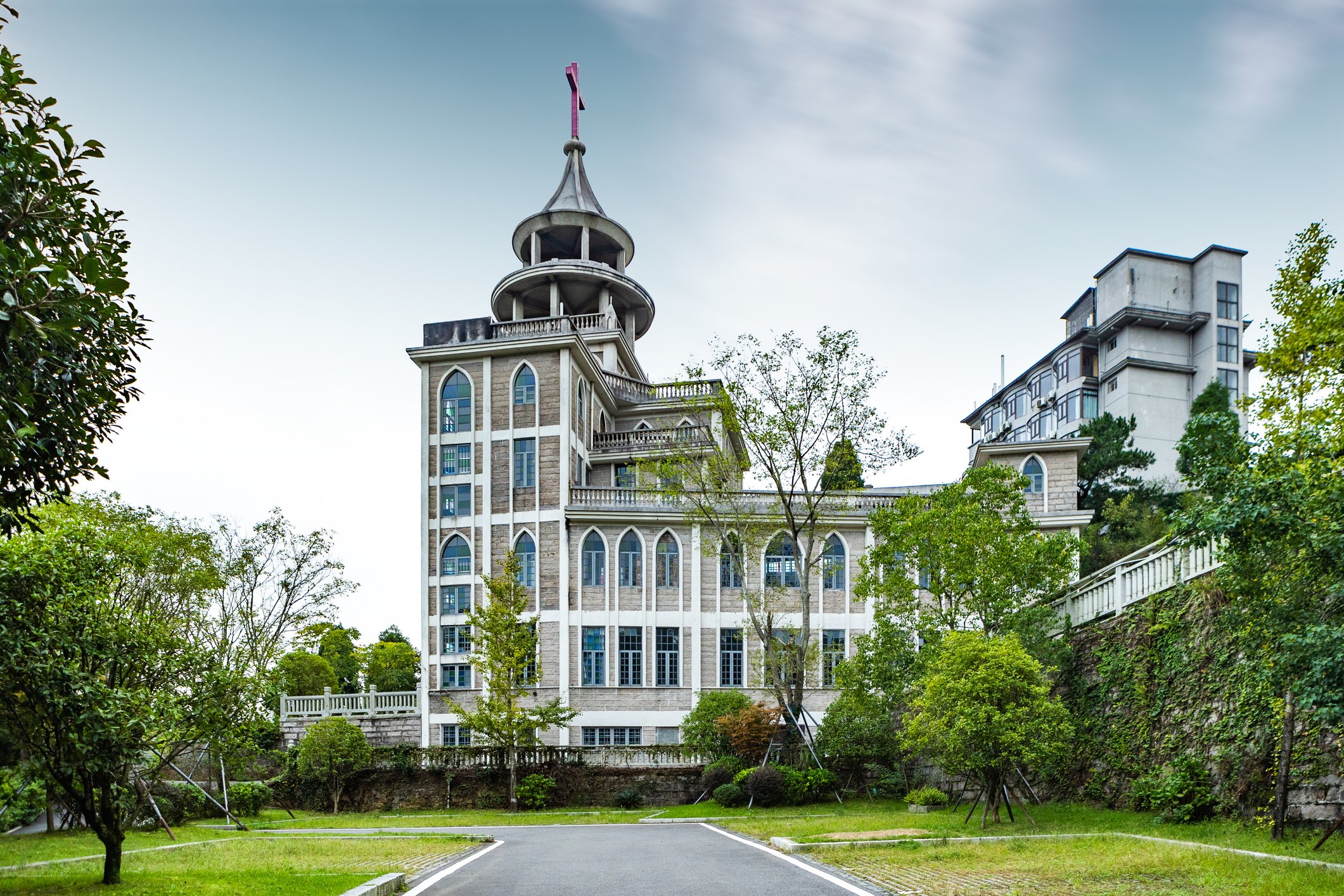 A large, ornate building with a tower featuring a pink cross at the top, surrounded by trees and greenery.