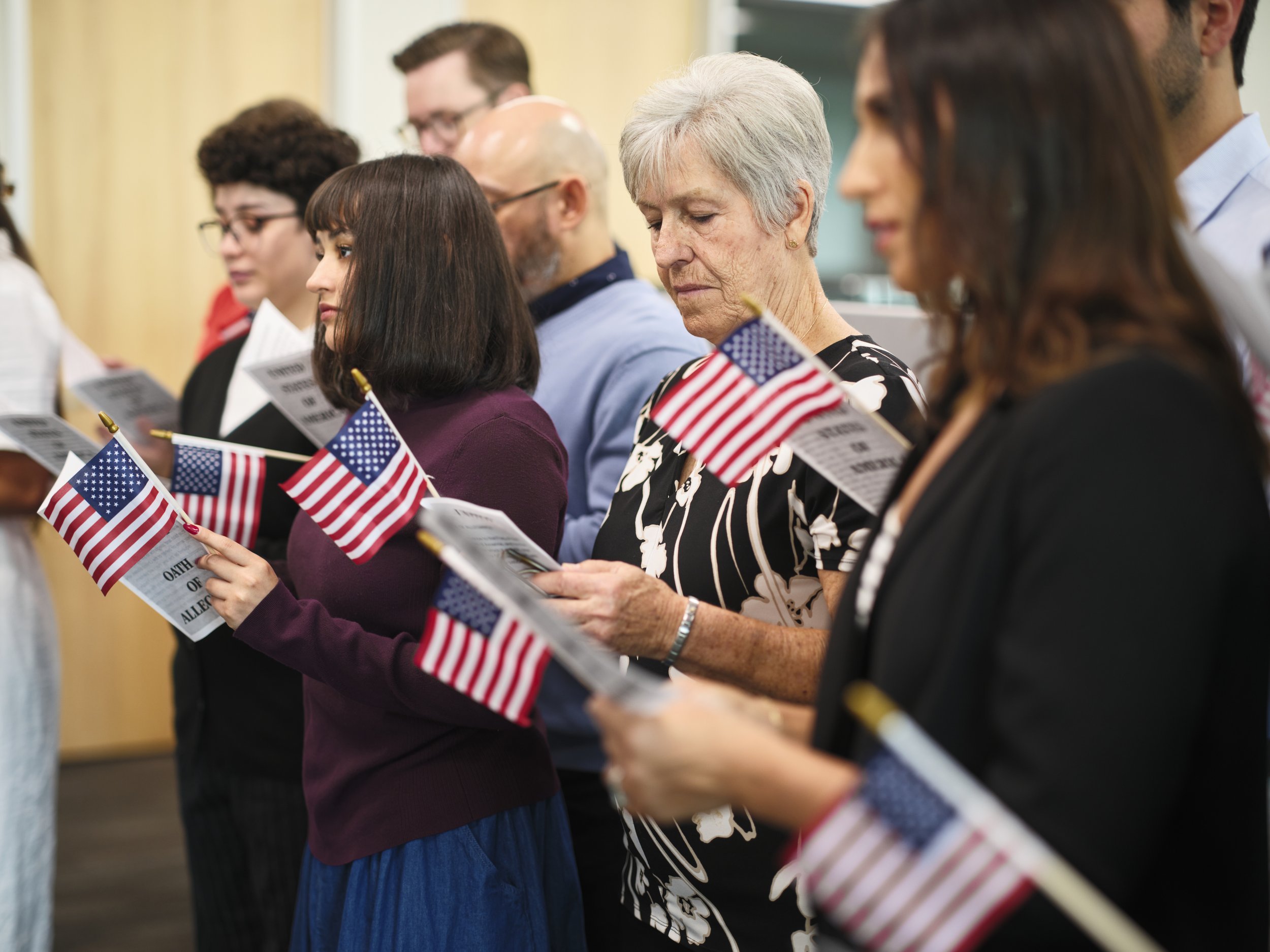 People attending a patriotic ceremony holding small American flags and reading documents, standing in a line indoors for the American citizenship swearing.
