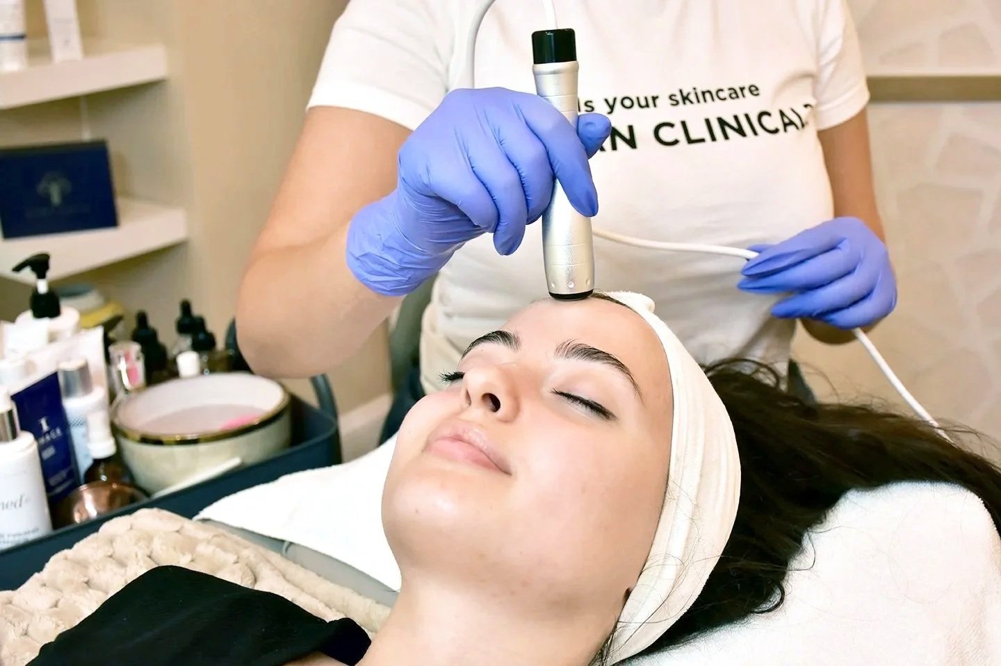 A woman receiving a facial treatment with a handheld device on her forehead at a skincare clinic, wearing a headband and relaxed with eyes closed.