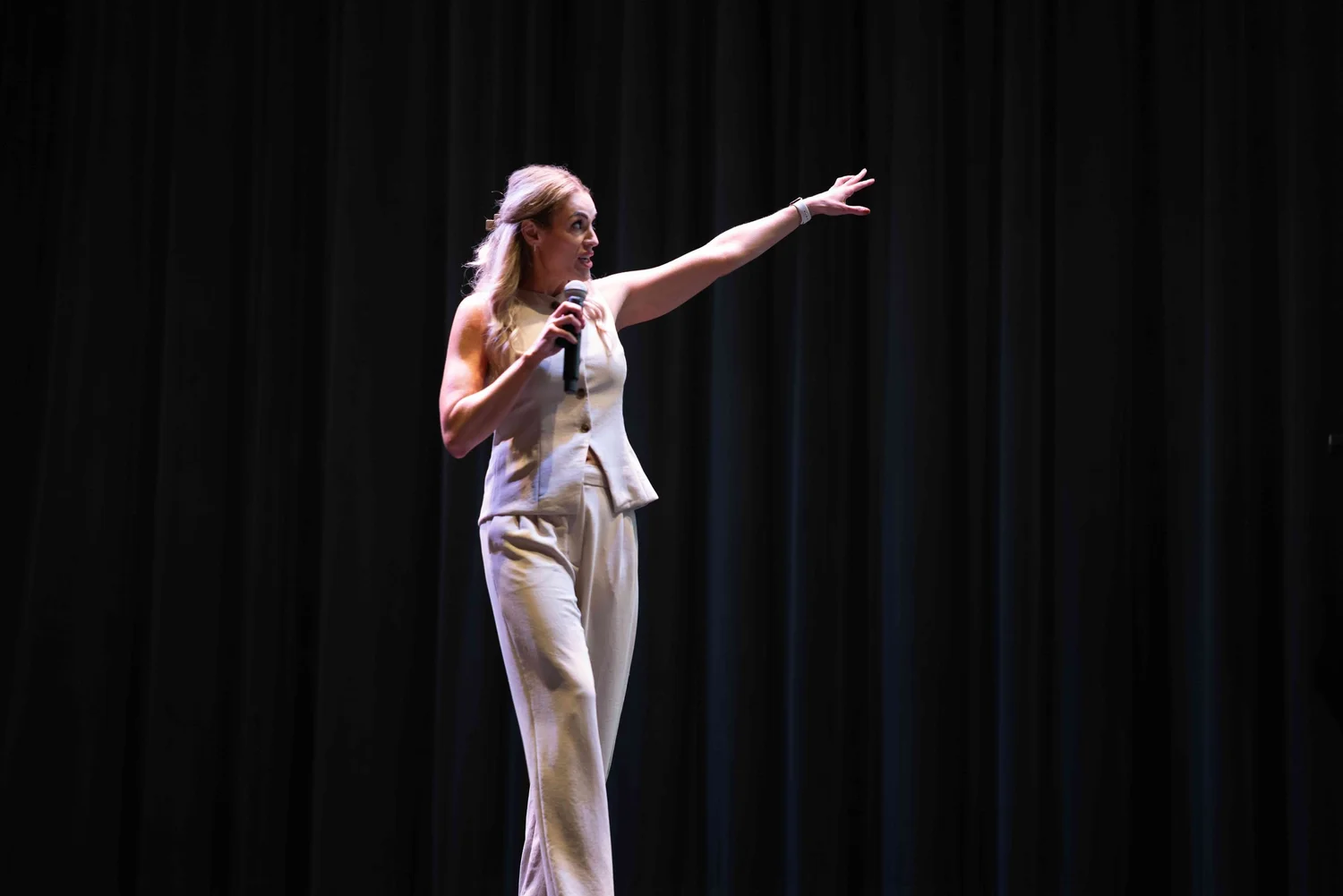 A woman in a beige outfit stands on a stage with black curtains, holding a microphone and pointing to the right.