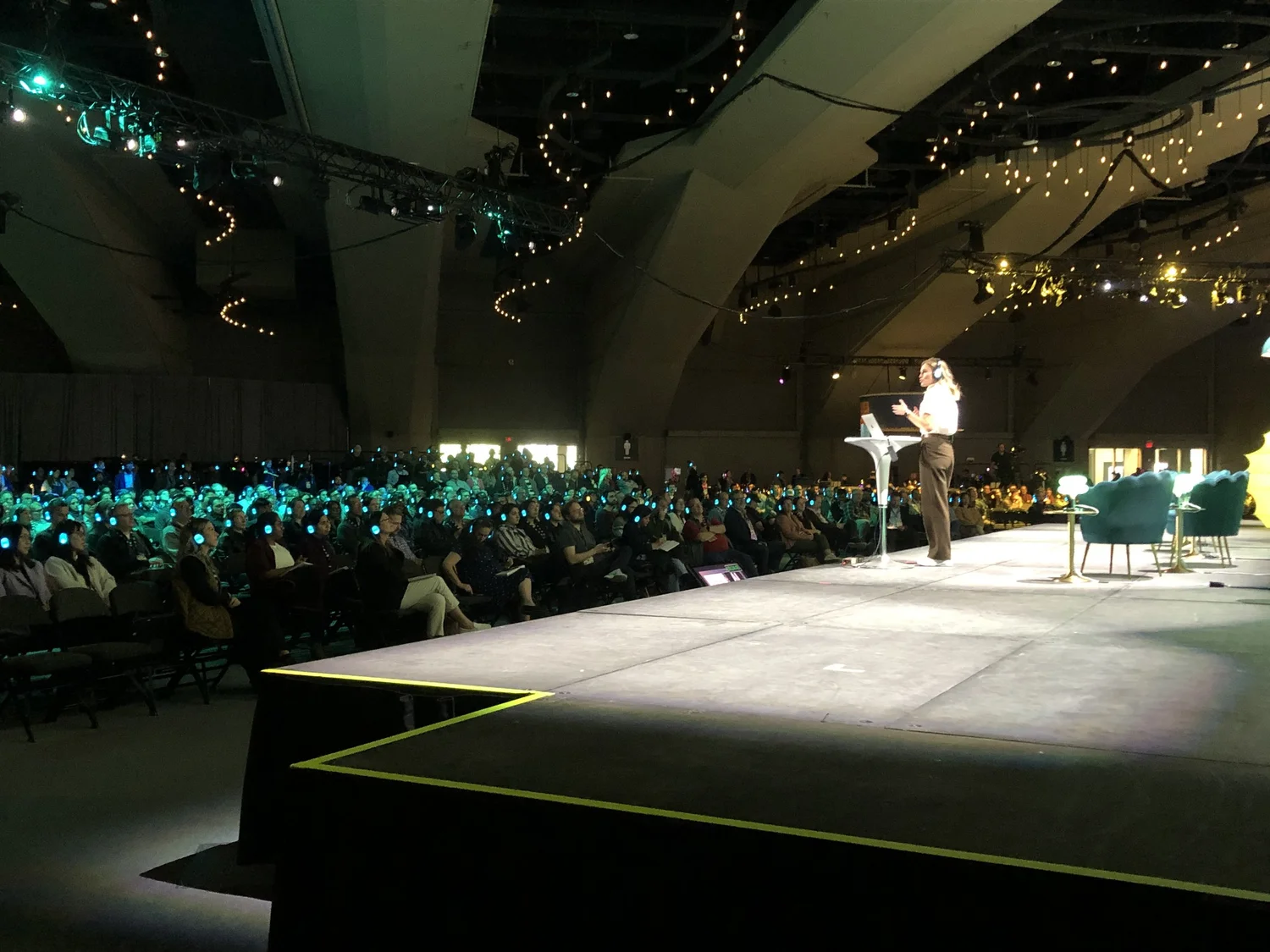 A woman giving a presentation on stage with a laptop and microphone, in front of a large seated audience in a conference hall decorated with string lights.