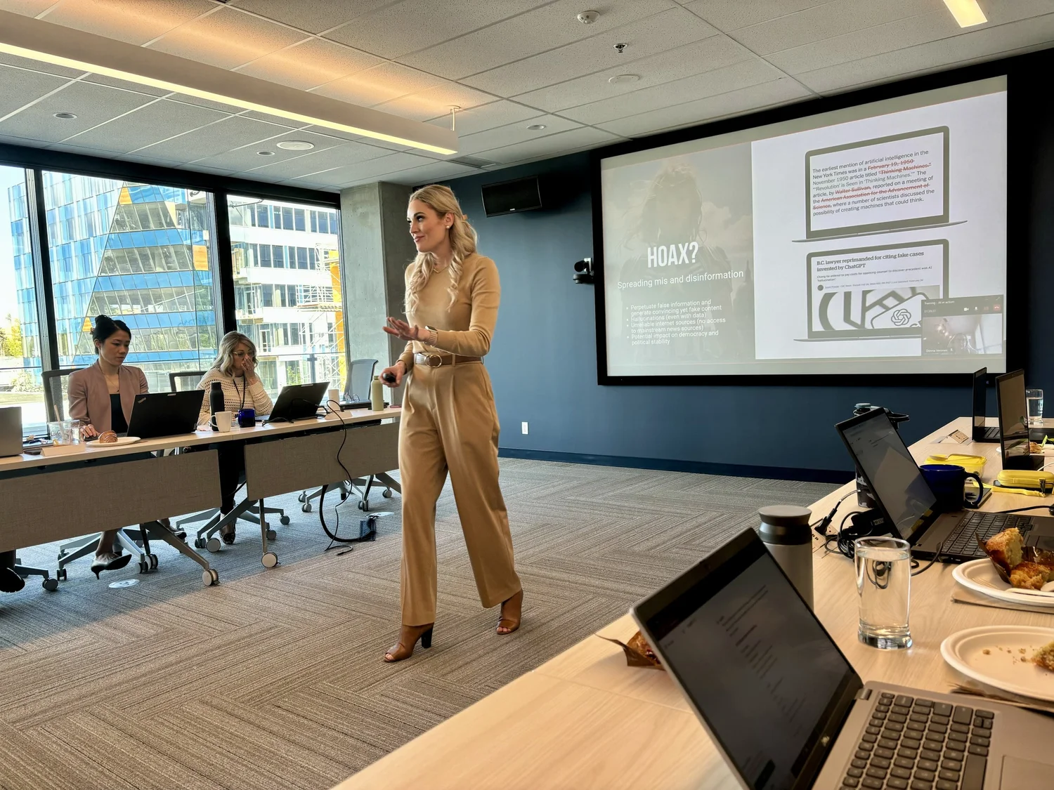 A woman giving a presentation in a conference room with large windows and city buildings outside. Three other women are seated at a long table with laptops, while a large screen displays a slide titled 'HOAX? Spreading mis and disinformation.' Plates with food, cups, notebooks, and glasses are on the table.