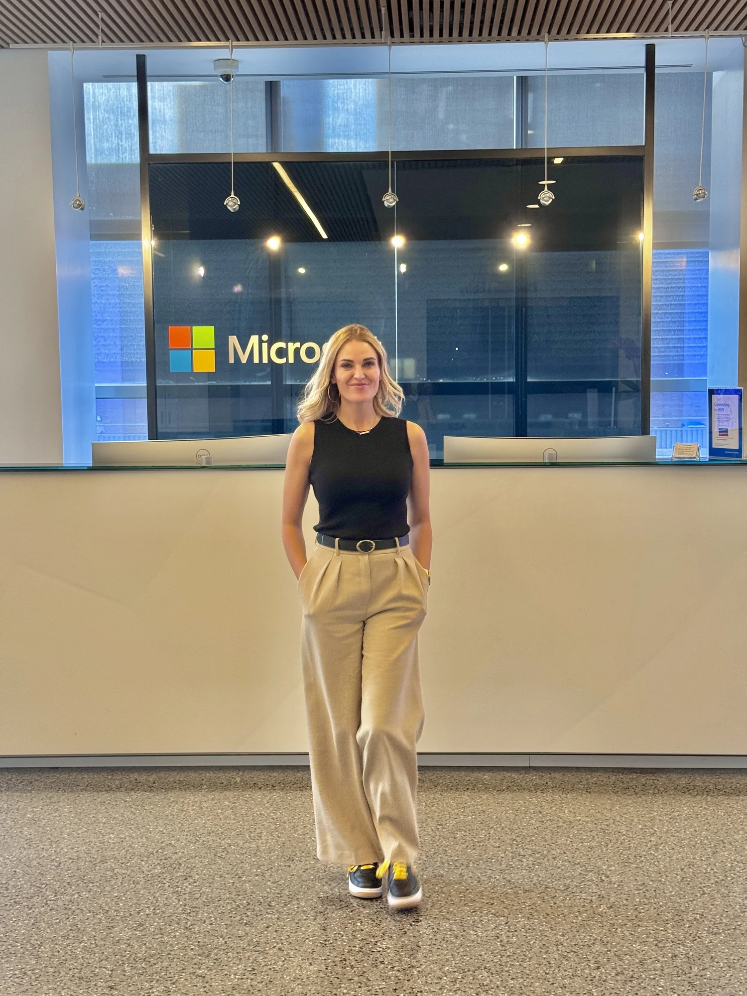 A woman with blonde hair, wearing a black sleeveless top, beige high-waisted pants, and black sneakers with yellow accents, standing in front of a reception desk at a Microsoft office.