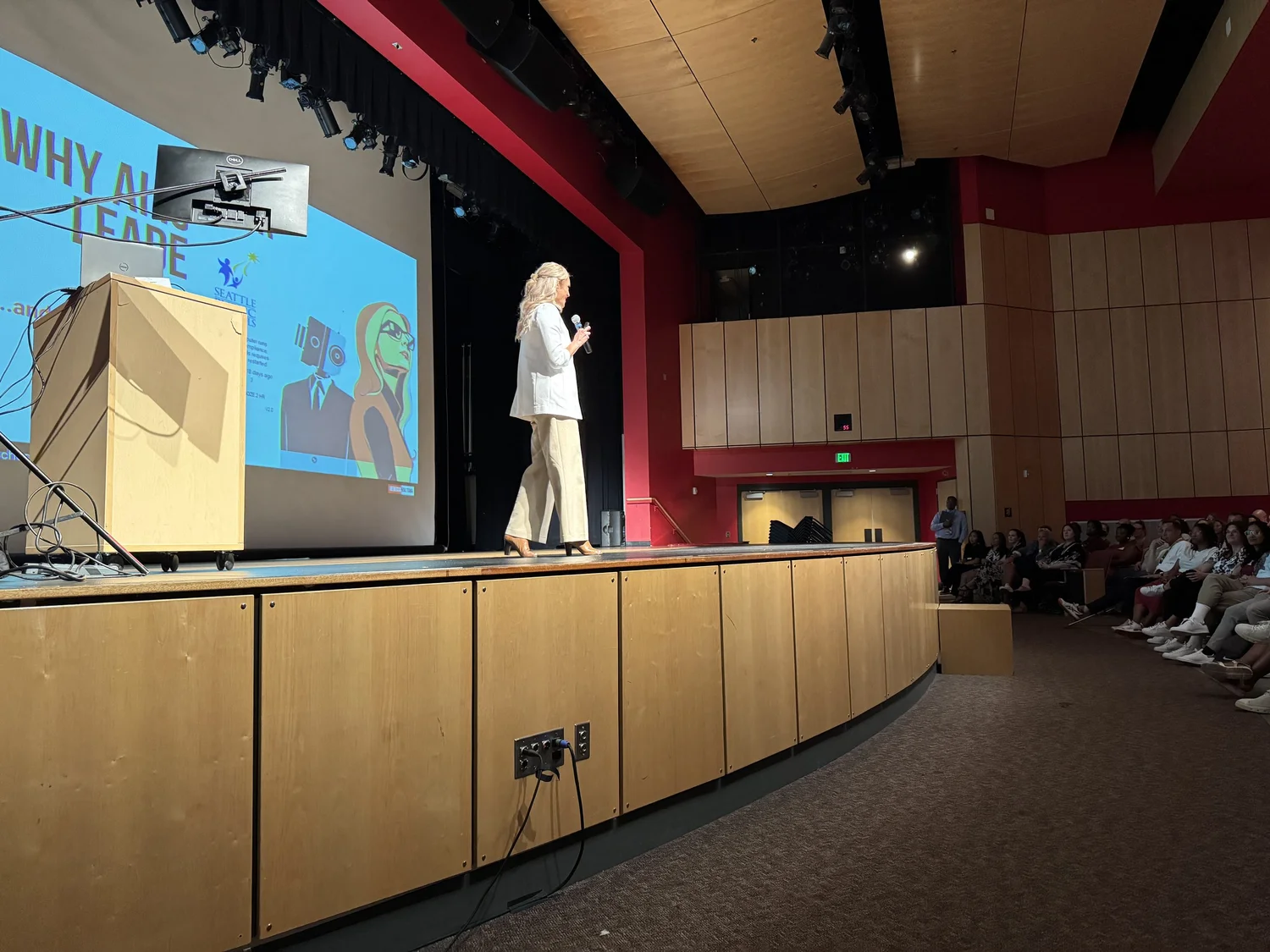 A woman with long, curly blonde hair is standing on a wooden stage, holding a microphone and giving a presentation in a large auditorium. A large screen behind her displays a slide with colorful illustrations and text, with part of the text reading 'WHY AL...LEAD'. The audience is seated on the right side of the image, and some audience members are looking towards the stage.