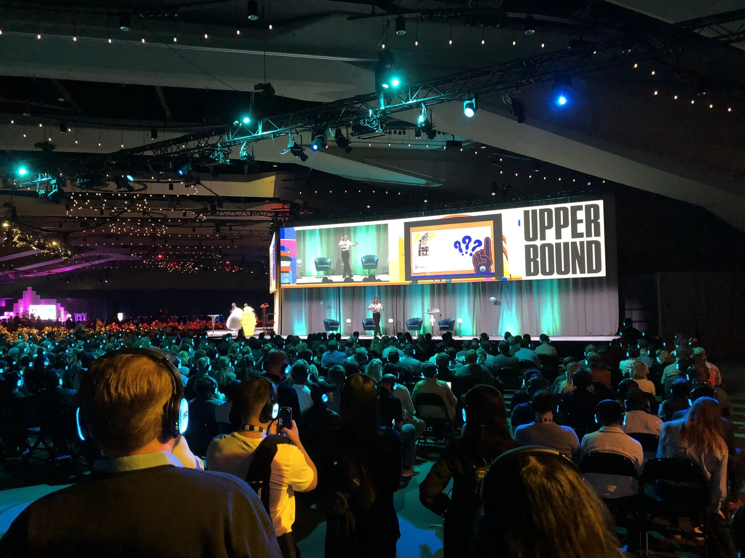 Audience attending a conference or presentation in a large indoor auditorium with a stage, large screen, and speakers, featuring a speaker on stage and various visuals, with colorful lighting and a dark ceiling.