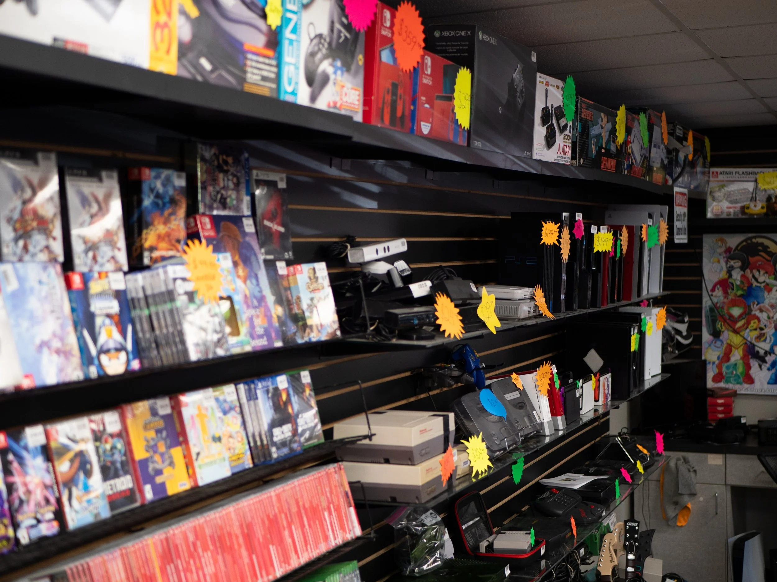 Shelf in a store filled with video games, gaming consoles, and accessories, with colorful sale tags attached.