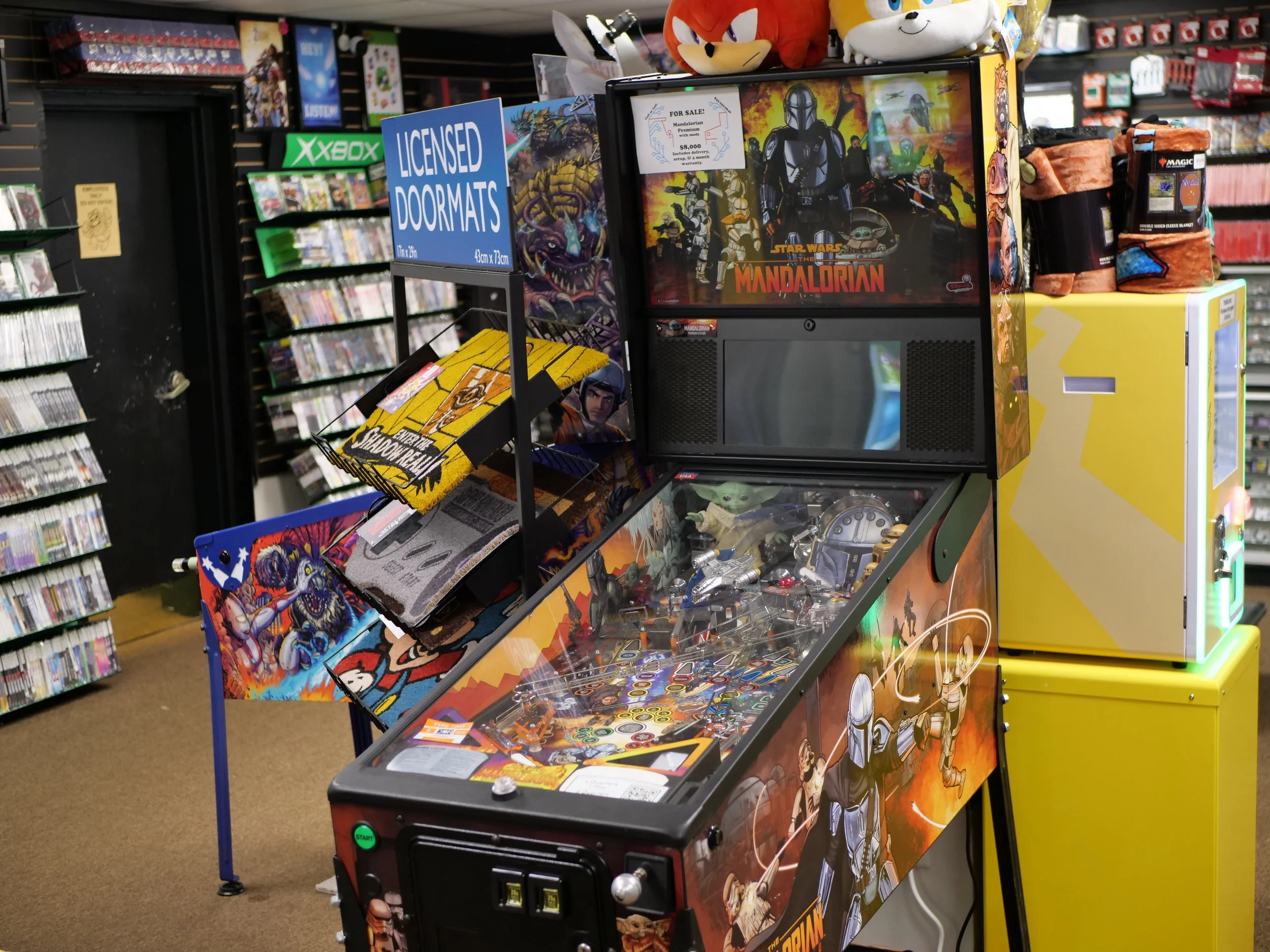 A Star Wars-themed pinball machine featuring characters from Star Wars, Mandalorian, and other sci-fi themes, with plush toys on top and sign for licensed door mats beside it in a video game store.