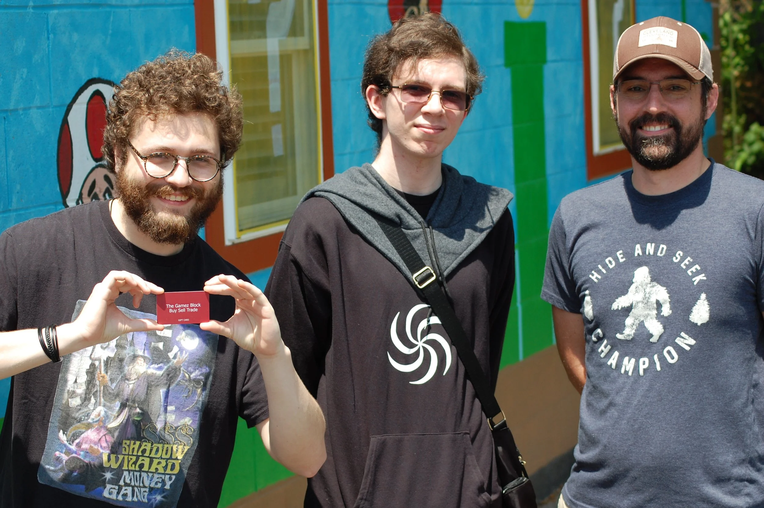 Three men standing outdoors in front of a colorful mural; the man on the left has curly hair and beard, holding a card; the middle man has straight hair and sunglasses, wearing a hoodie; the man on the right has a beard and is wearing a T-shirt and c