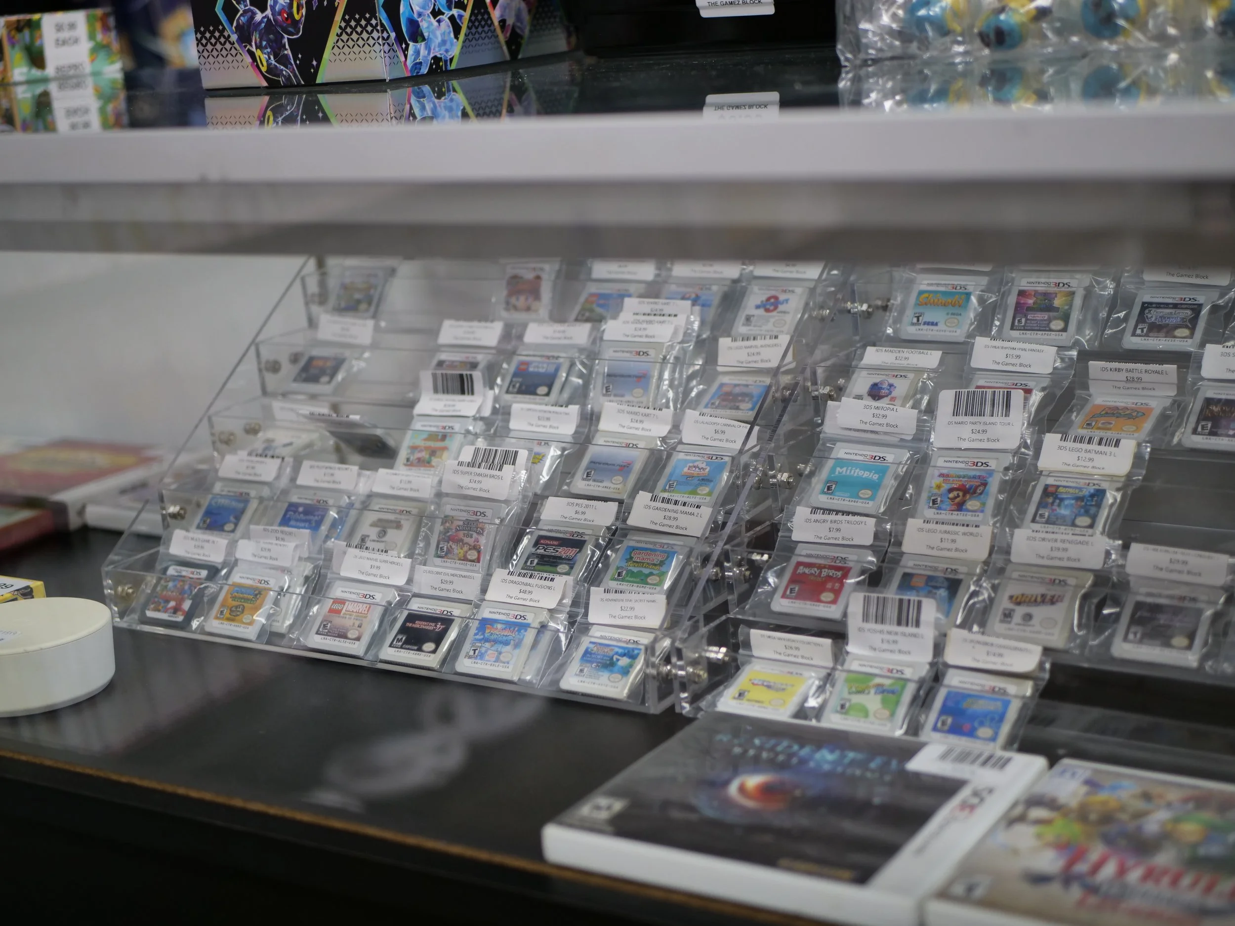 Display case with multiple Nintendo 3DS game cartridges in clear plastic holders, labeled with prices. There are game boxes on top and some on the table. Overall, a video game store setup.