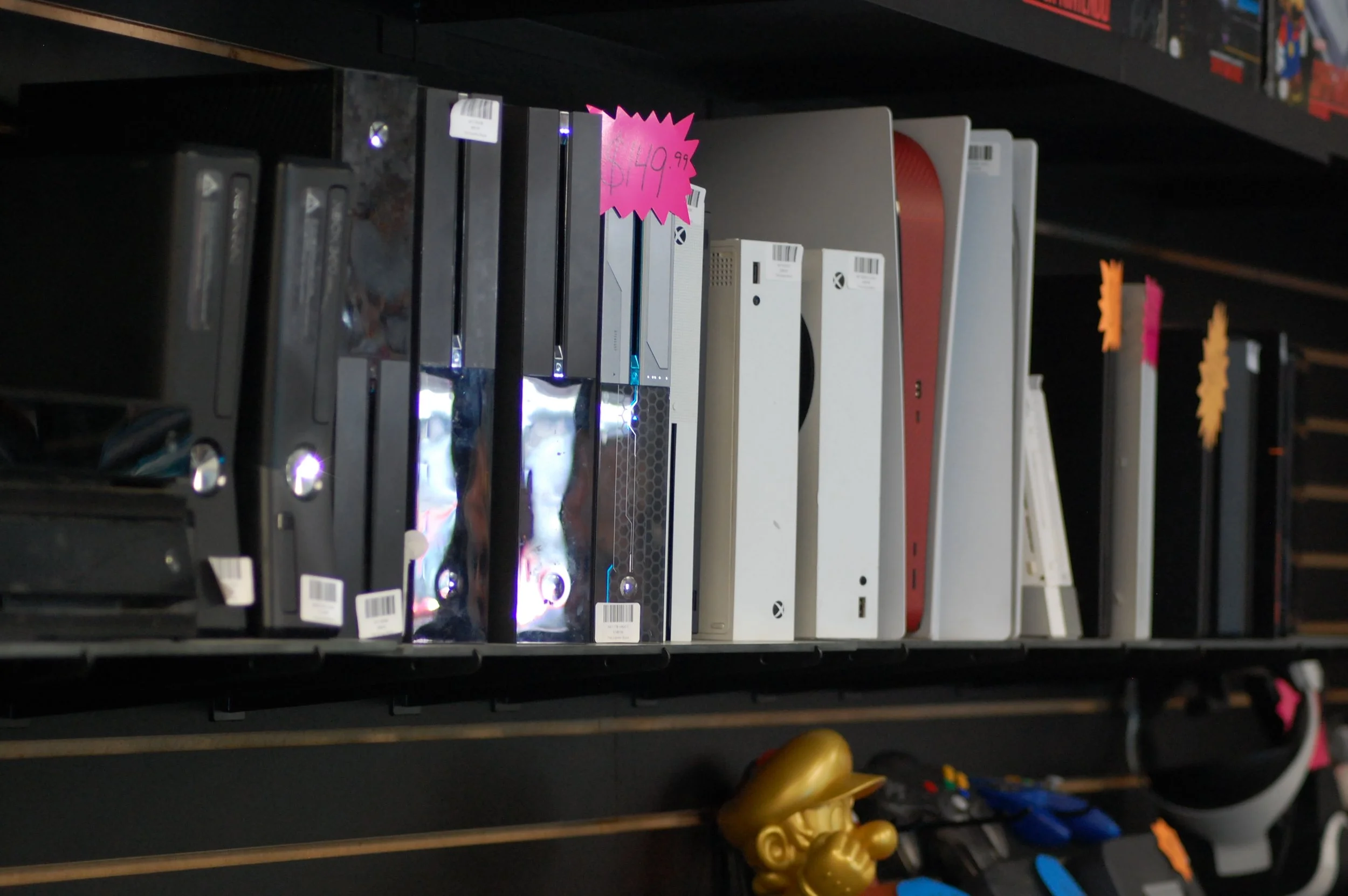 Video game consoles on a store shelf, including Xbox Series X models, with a pink price tag reading $149.99, and some gaming controllers below.