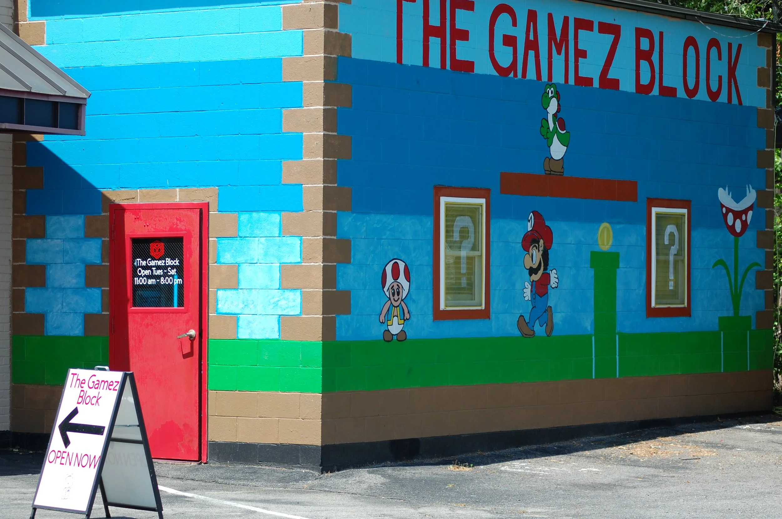 Colorful mural on a building exterior featuring characters from Super Mario, including Mario, Luigi, Toad, Yoshi, and Flower, with red and blue backgrounds and the words 'The Gamez Block' painted at the top. A red door with a black sign and a white s