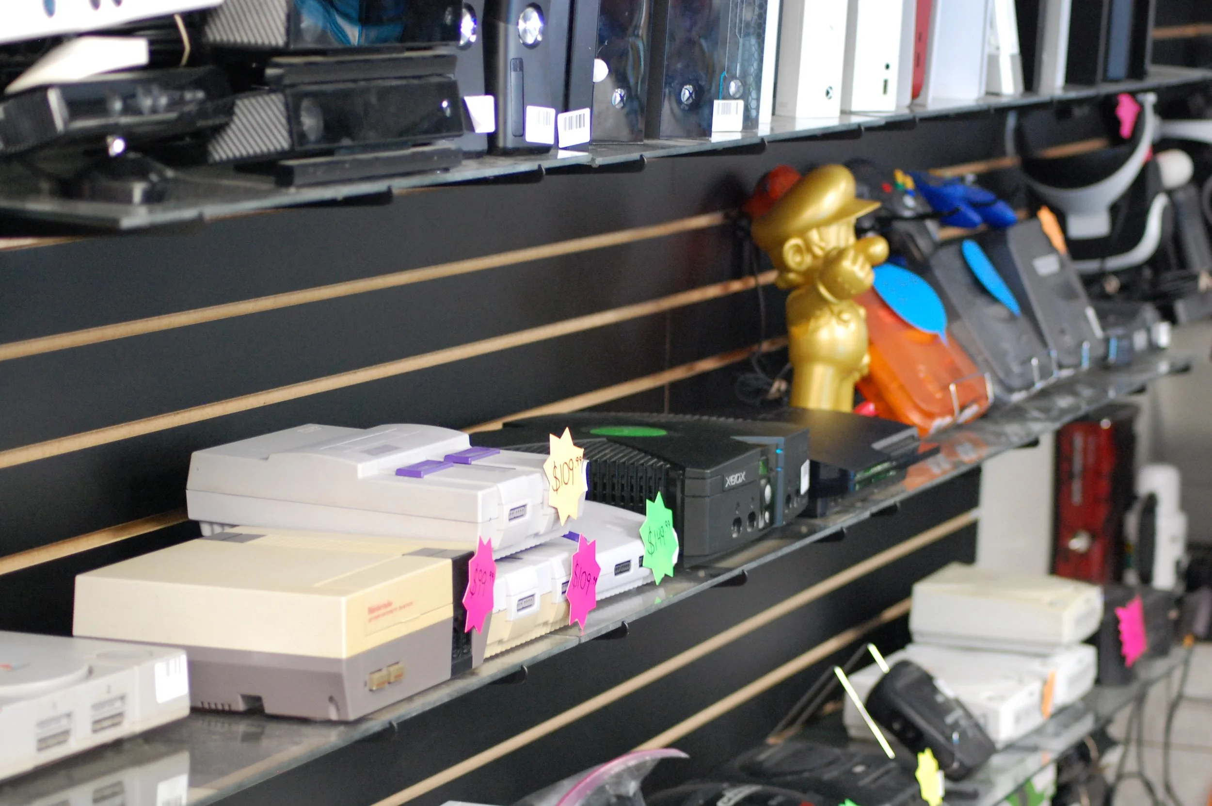 A black shelf with vintage gaming consoles, a golden Mario statue, and various electronic items with colorful sale tags.