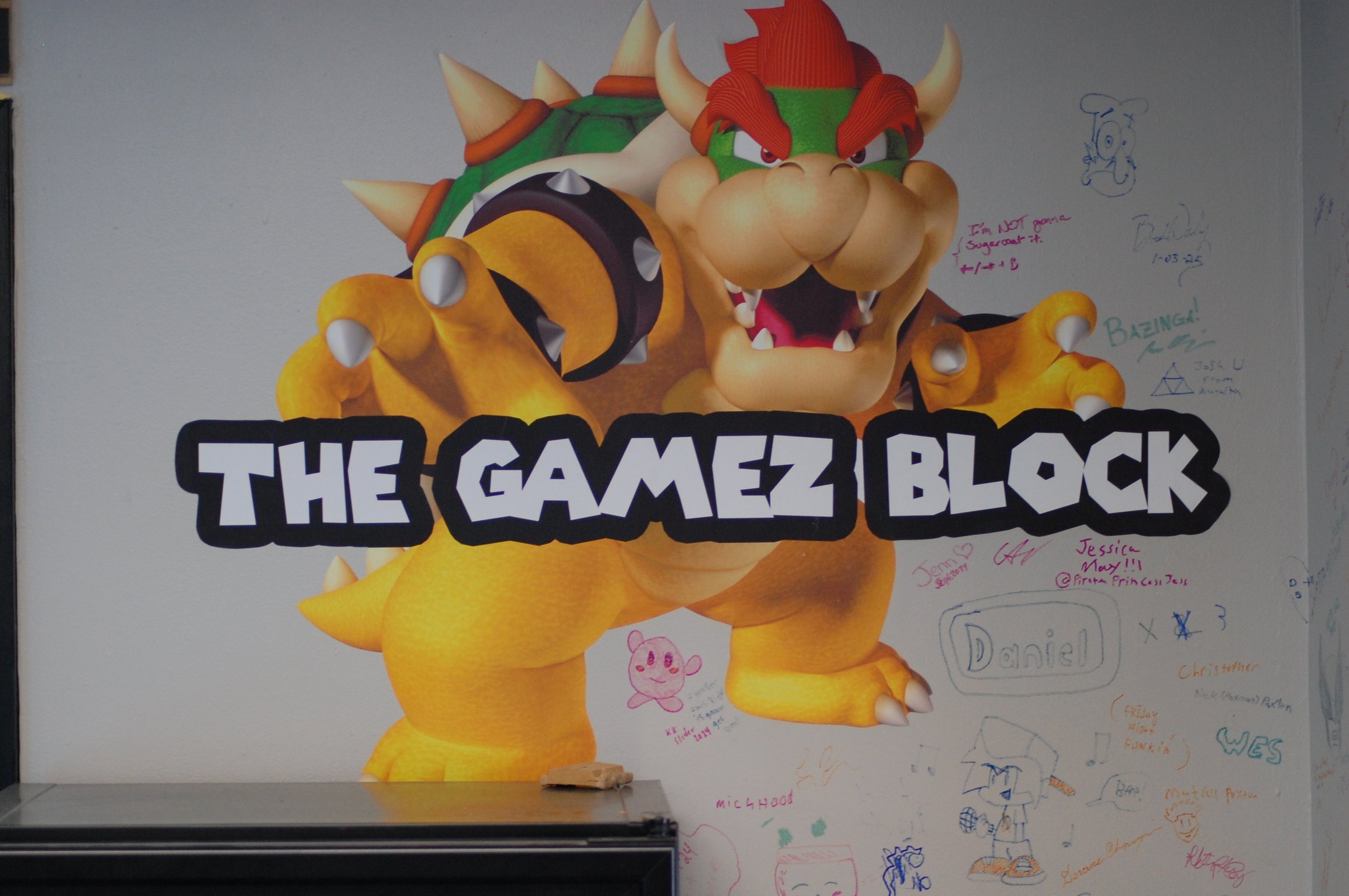 A large poster featuring Bowser from Super Mario with the text 'The Gamez Block.' The poster has various handwritten notes and doodles around it, some mentioning names, characters, and messages.
