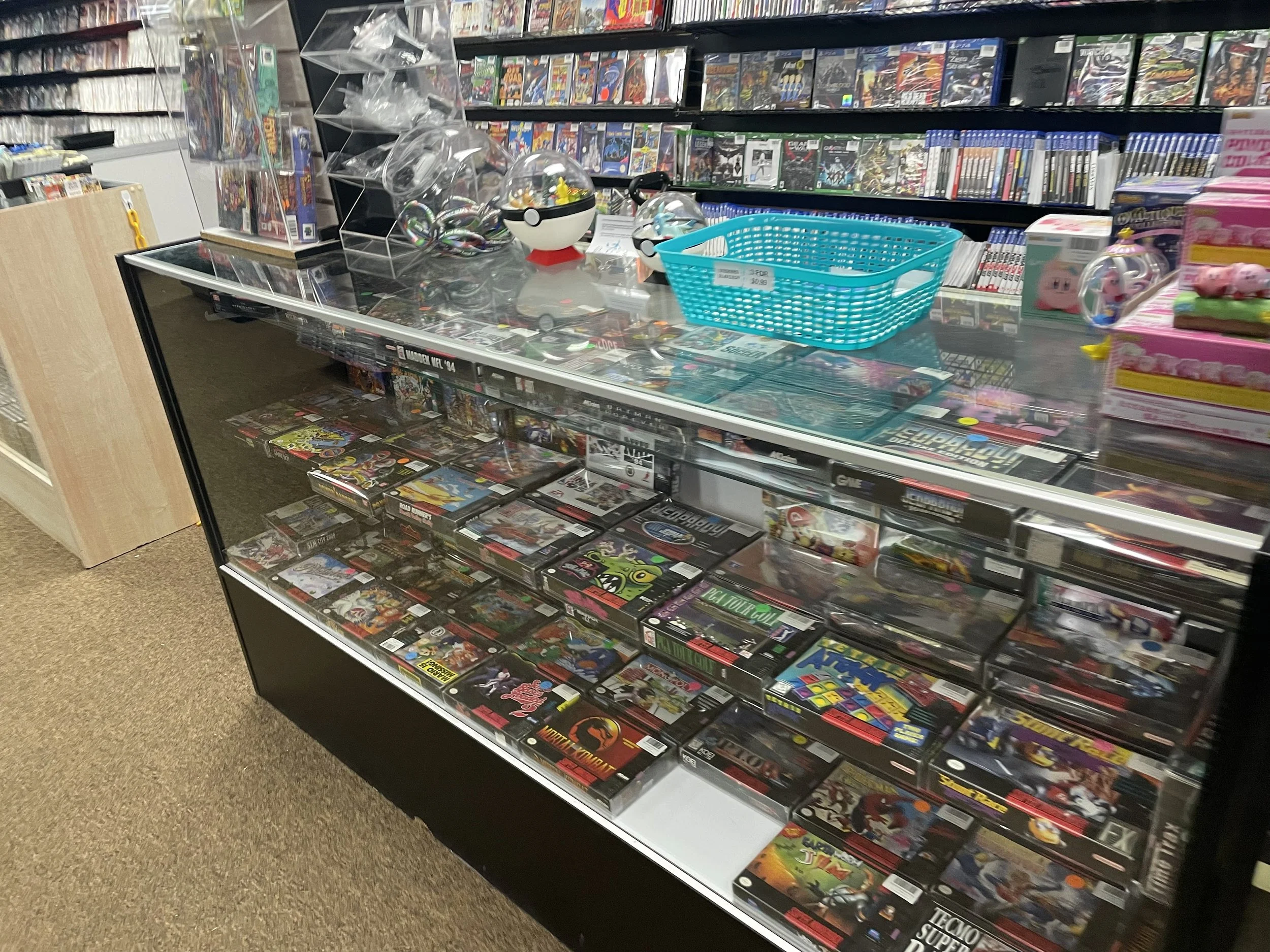 Display case filled with various video game cases in a store, with shelves of video game boxes behind it, and some collectible toys on top.
