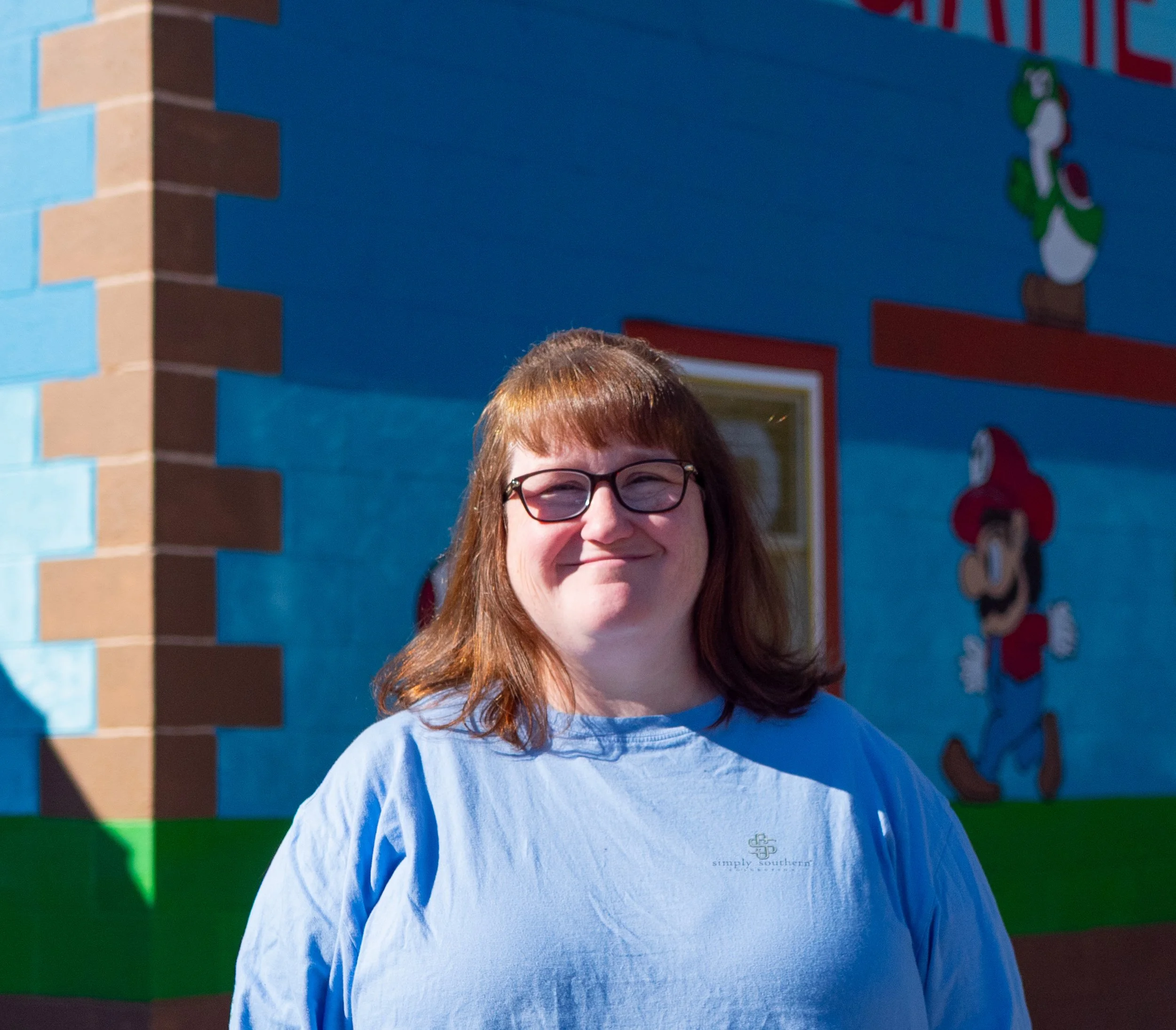 A woman with shoulder-length reddish brown hair and glasses smiling outdoors in front of a colorful mural featuring characters like Luigi and Yoshi from Mario.