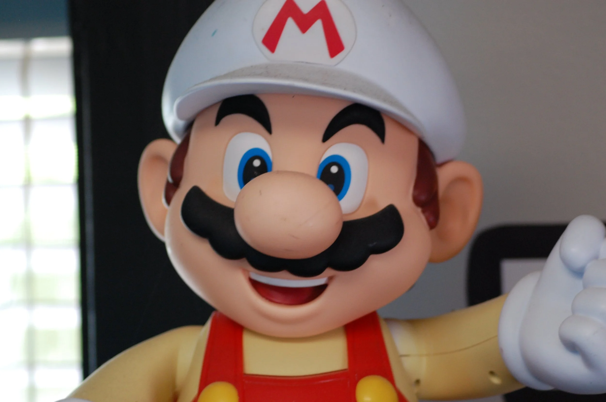 Close-up of a large Mario mascot statue with a white cap featuring a red 'M', a big nose, thick black mustache, blue eyes, and a red and yellow outfit, giving a thumbs-up gesture.