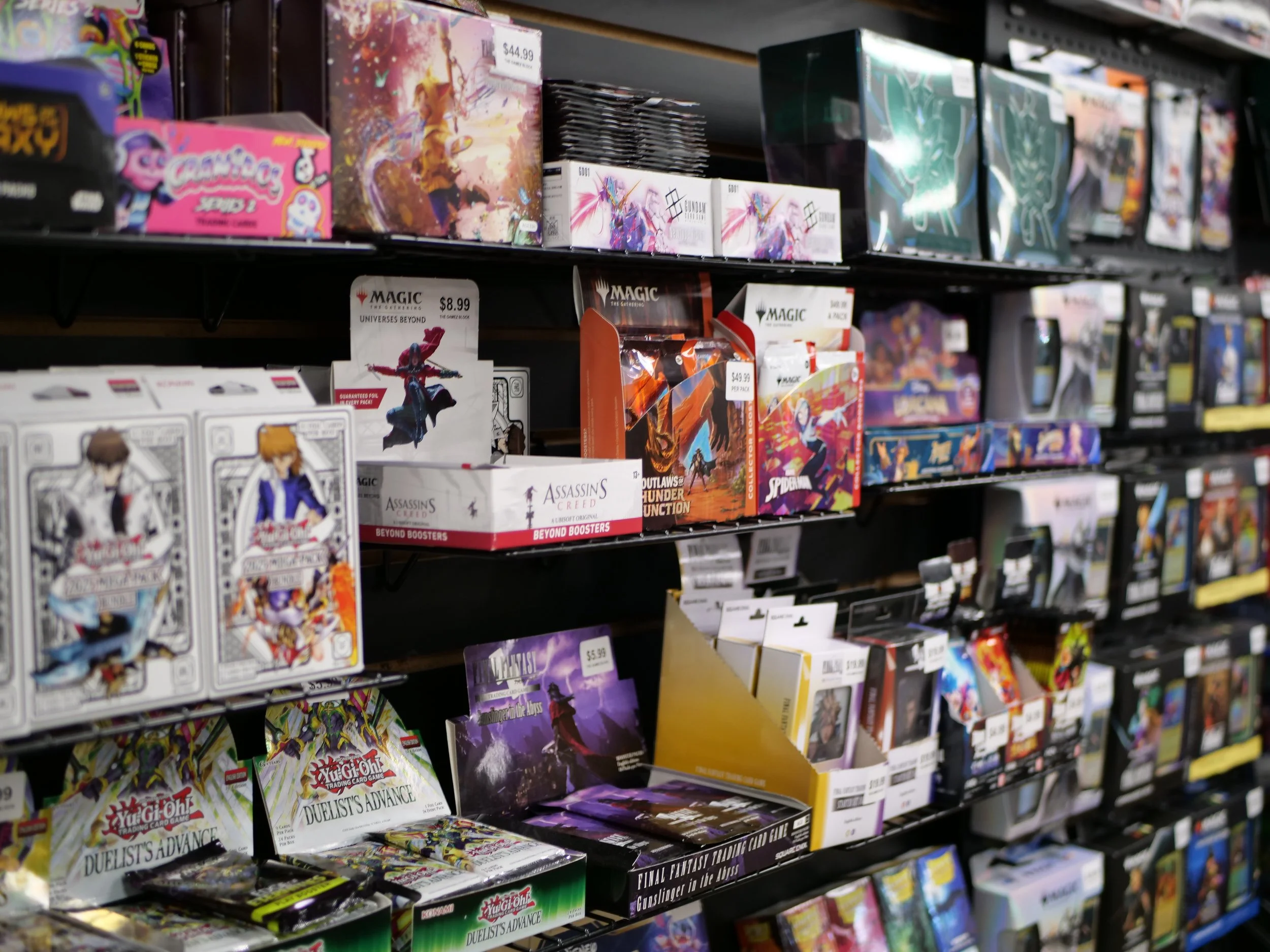 Video game card packs and booster boxes on store shelves, including Yu-Gi-Oh! Duelist's Advance and Magic the Gathering products.