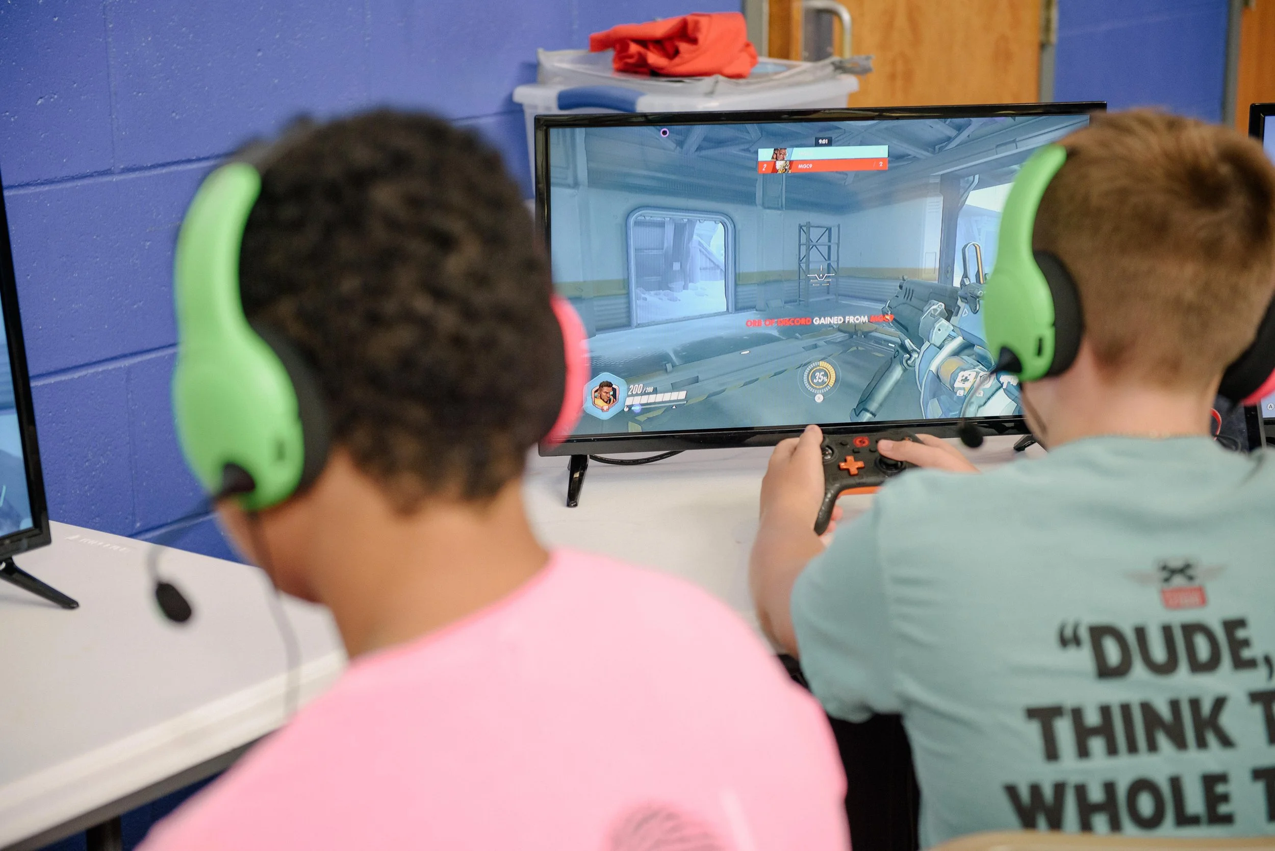 Two boys playing a first-person shooter video game on a computer monitor, wearing green and black gaming headsets, in a gaming room with a blue wall.