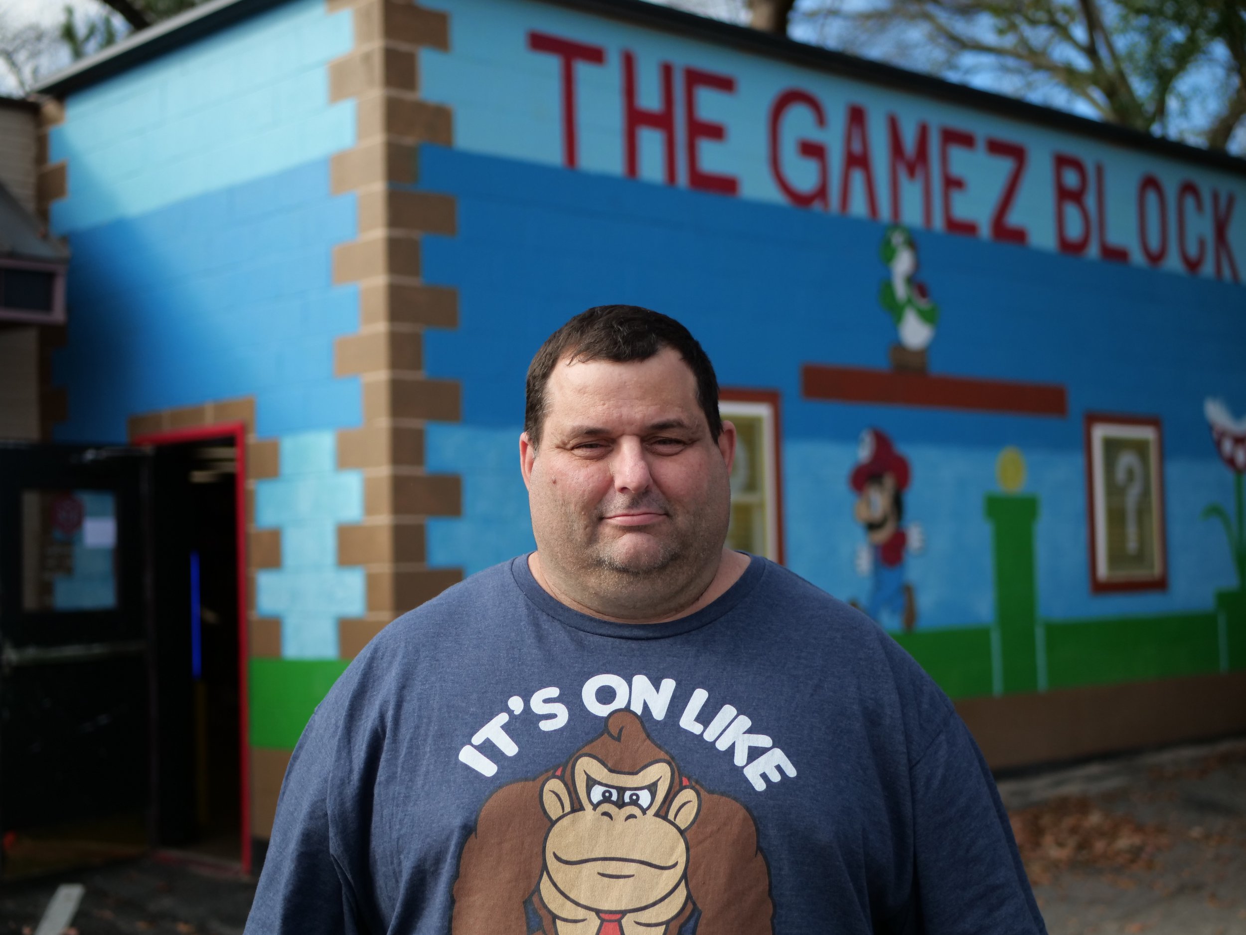 A man standing outdoors in front of a colorful mural wall with the words 'The Gamez Block' at the top. The man is wearing a blue t-shirt with a cartoon of a gorilla and the text 'It's on like.'