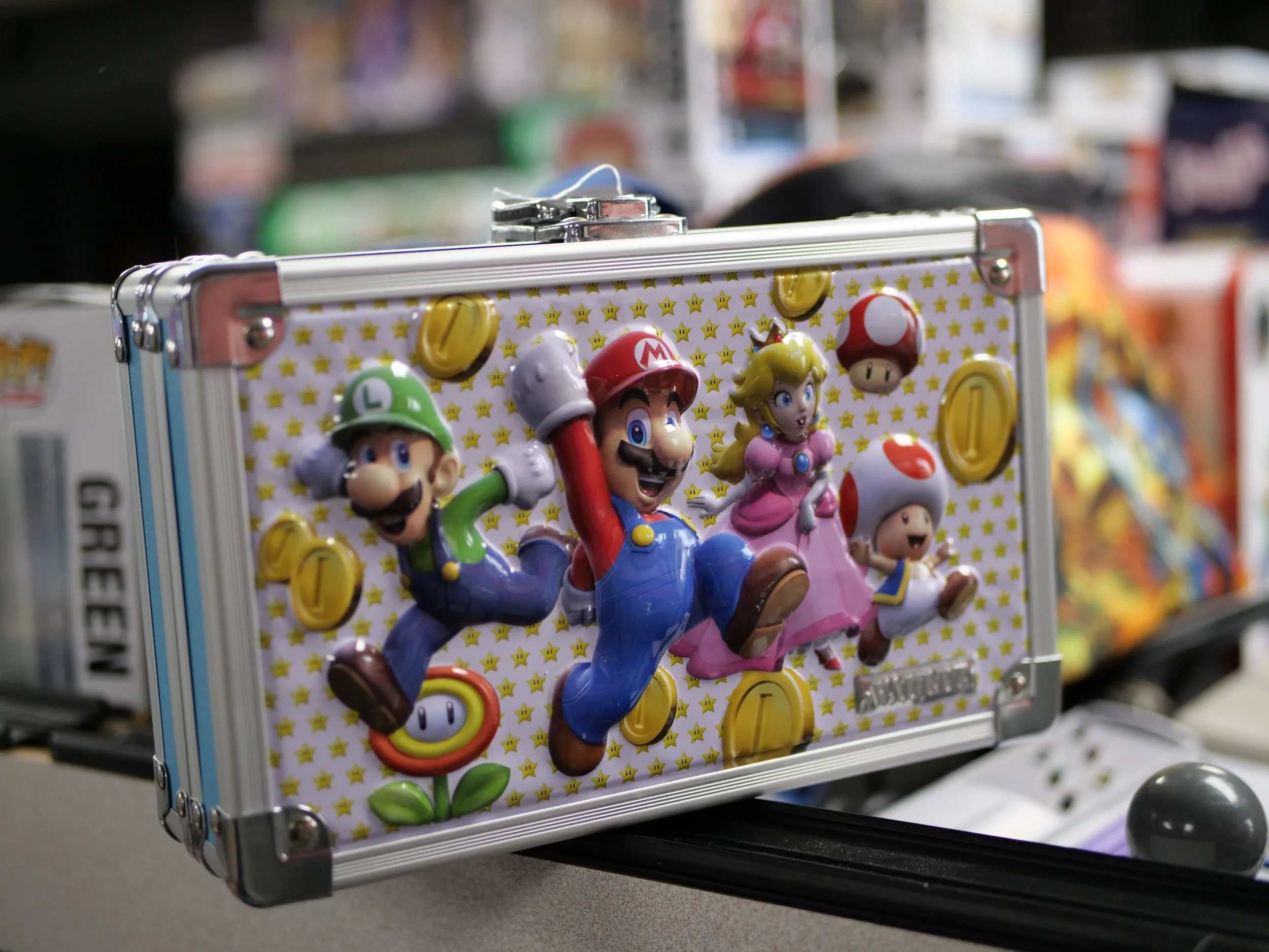 A metal box featuring 3D figures of Mario, Luigi, Princess Peach, Toad, and Toadette from Super Mario, surrounded by yellow coins and star patterned background.