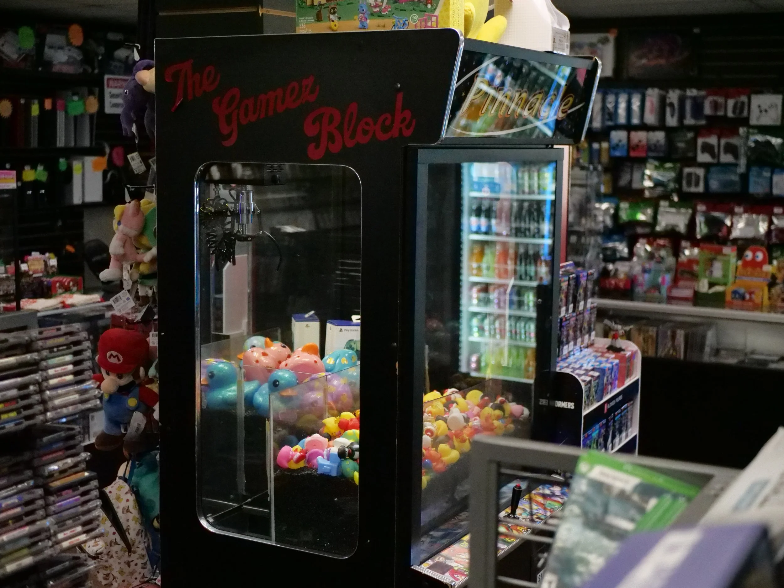 A claw machine filled with colorful toys and capsules inside a store with shelves of DVDs, games, and toys in the background.