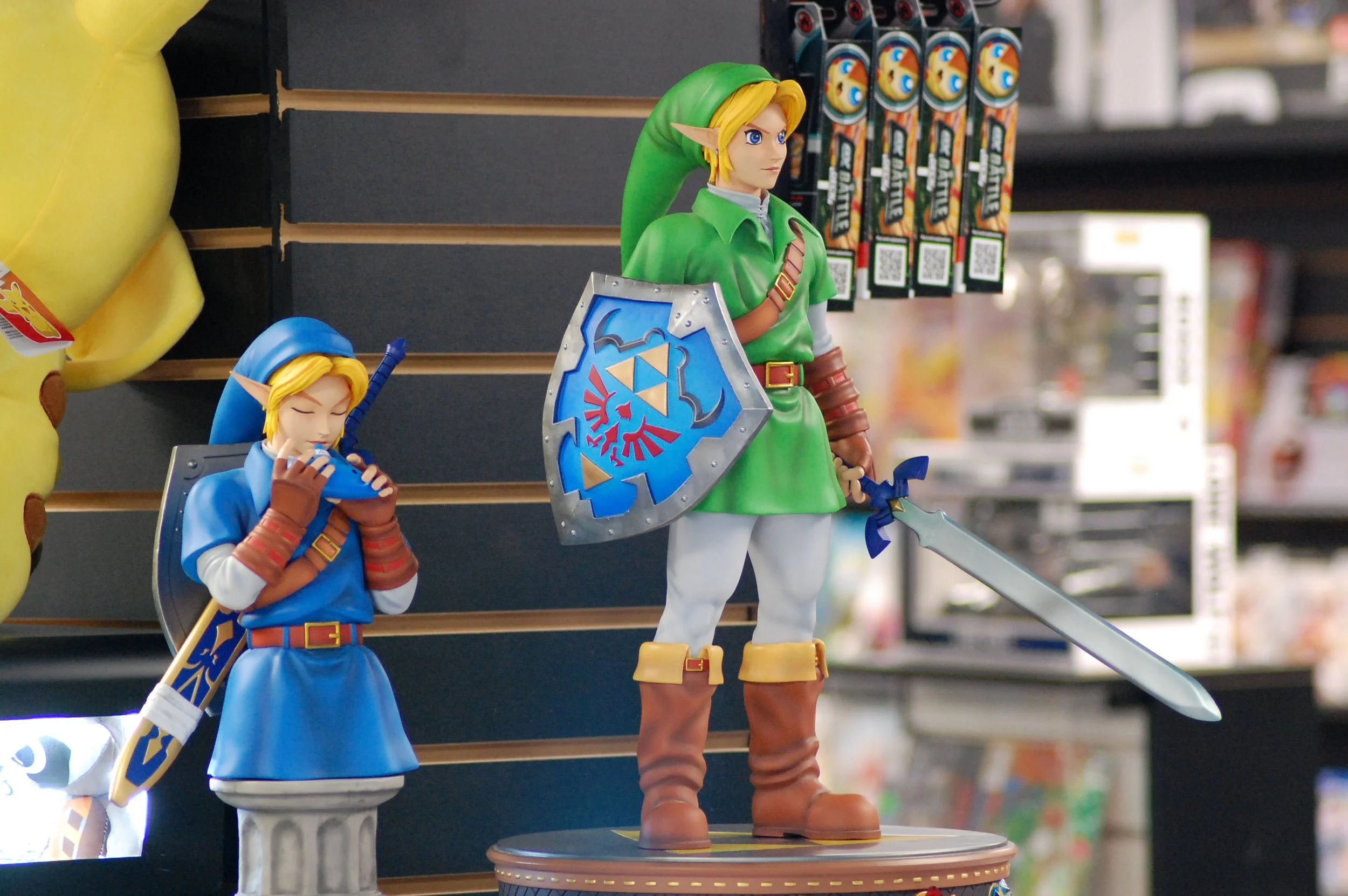 Figures of Link from The Legend of Zelda in two different poses, one standing with a sword and shield and the other with eyes closed holding a small item, displayed on shelves in a store.