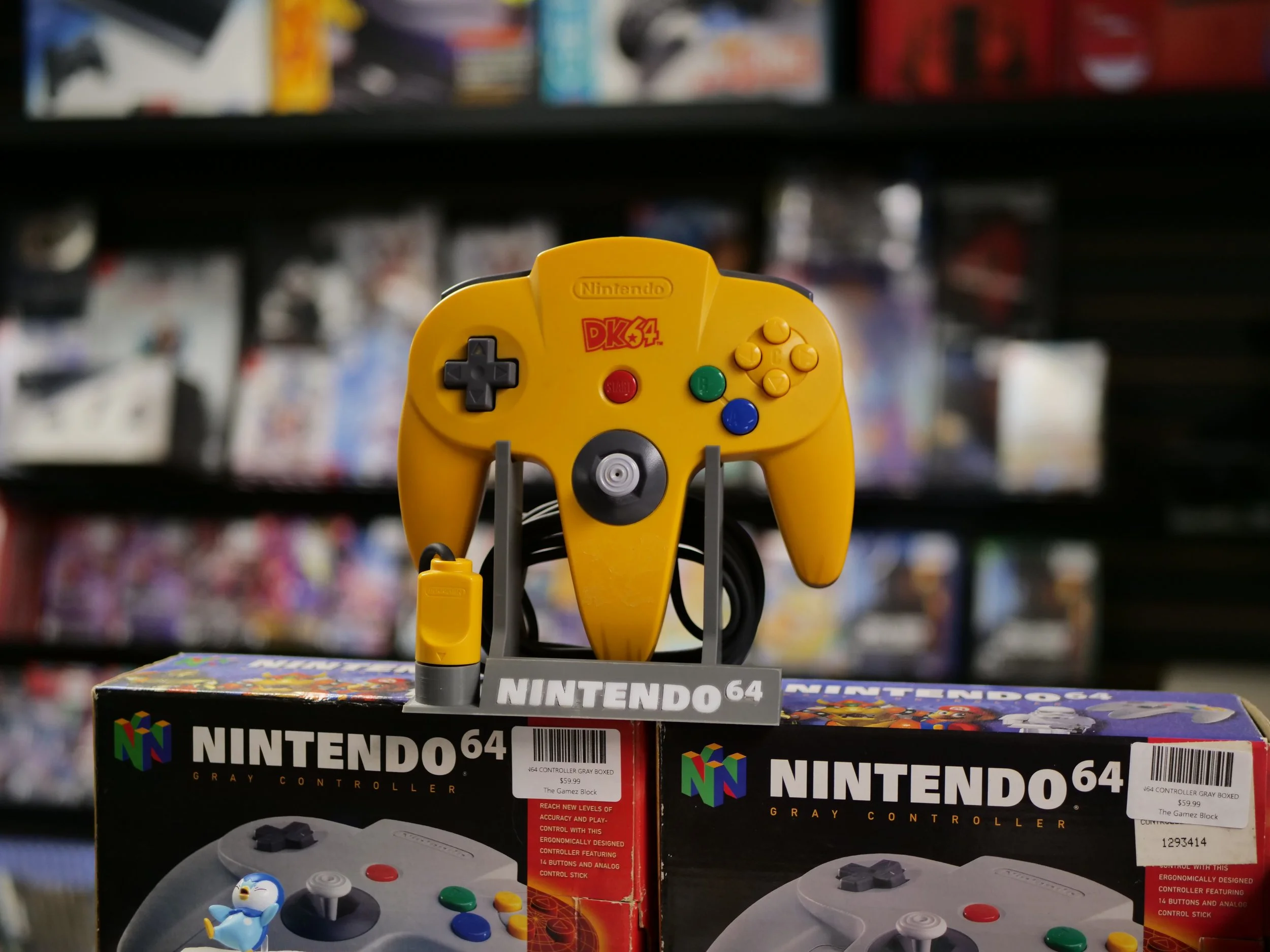 A yellow Nintendo 64 game controller on top of boxed Nintendo 64 controllers, with a background of video game shelves.