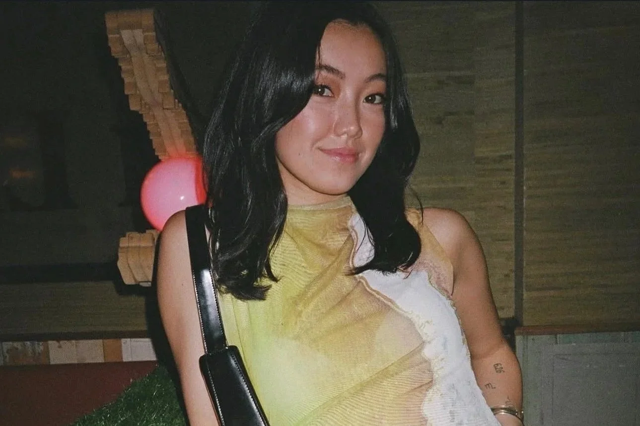 A young woman with shoulder-length black hair, wearing a sleeveless yellow and white top, poses indoors with a slight smile. There is a wooden wall and a pink balloon in the background.