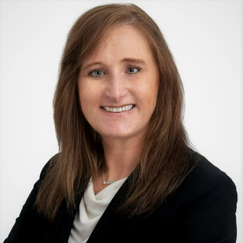 Professional headshot of a woman with shoulder-length brown hair, smiling, wearing a black blazer over a white top, against a plain light background.