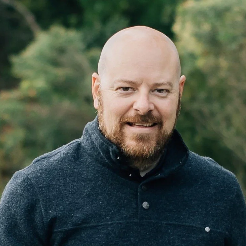 A smiling bald man with a beard outdoors, wearing a dark sweater, with green foliage in the background.