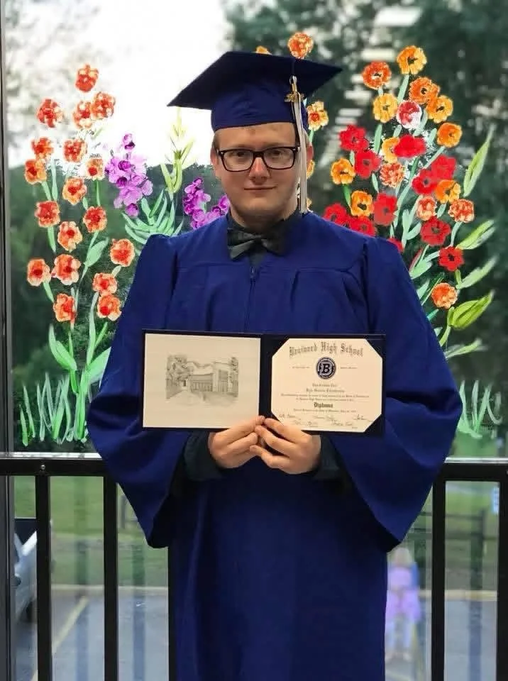 A young man in a blue graduation gown and cap holding a diploma, standing in front of a colorful floral background.