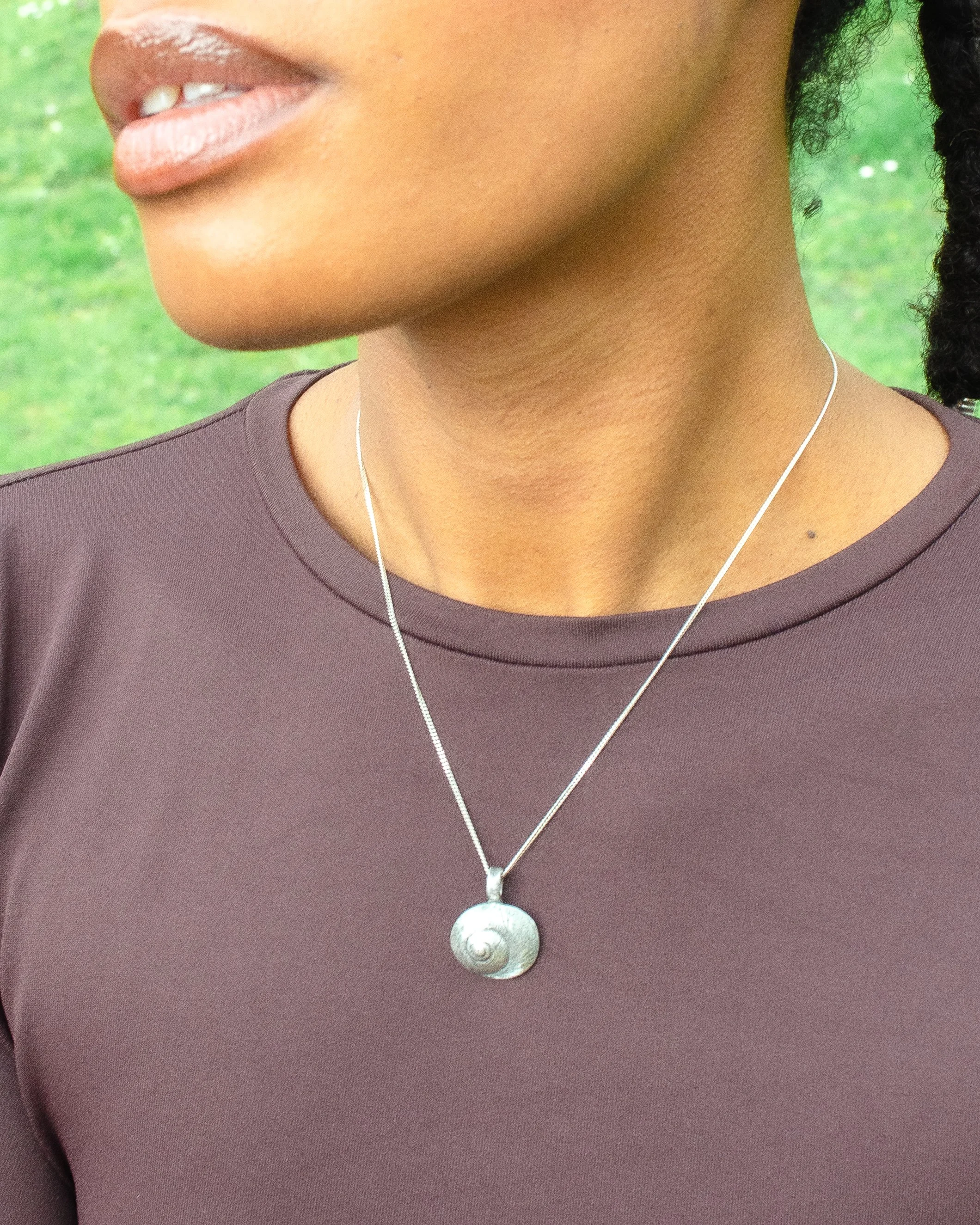 Fine silver round shell pendant on a silver chain, worn against bare skin, with a large white scallop shell in the background