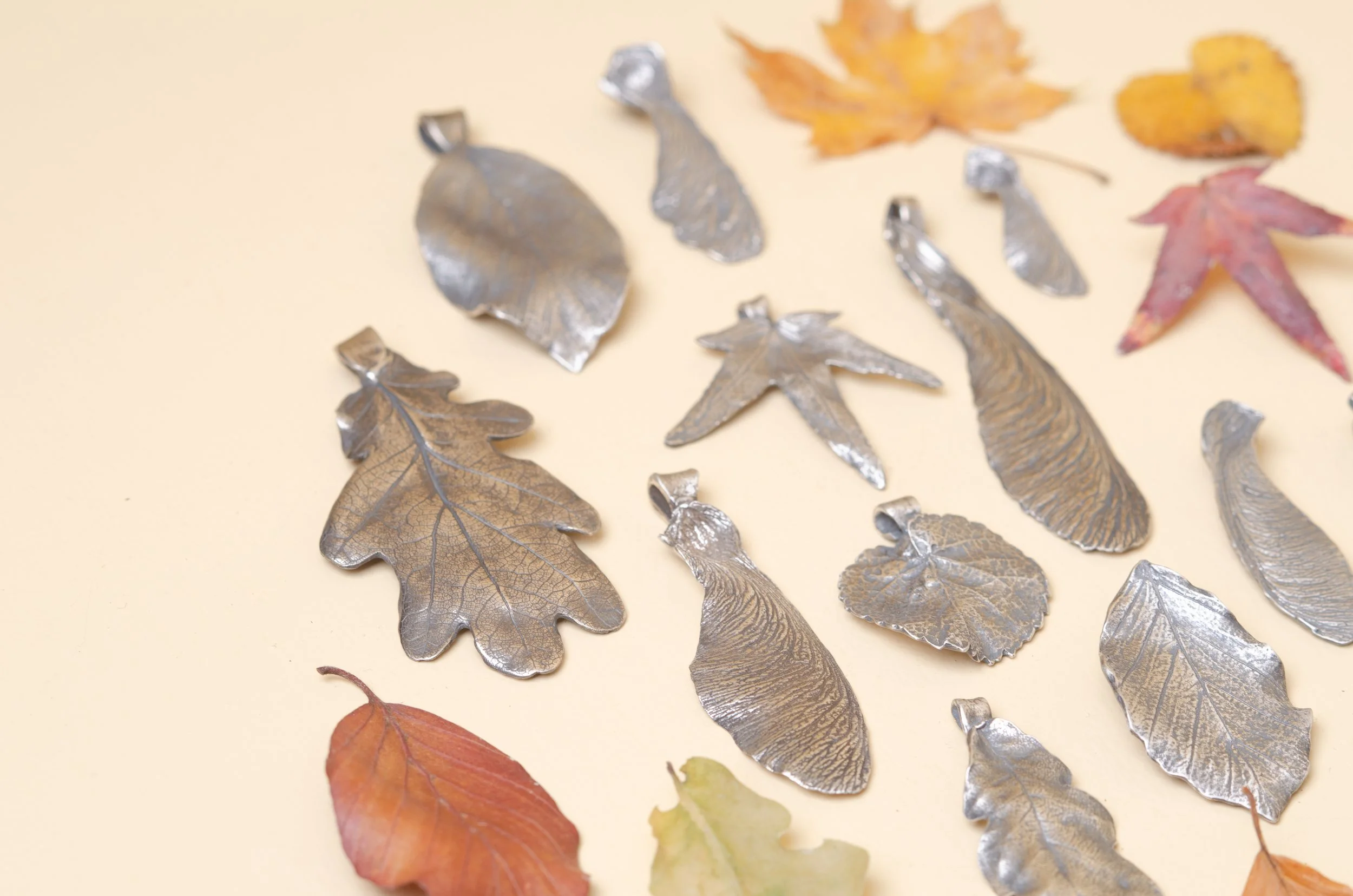 An artistic top-down view of various handcrafted sterling silver charms shaped like maple seeds and oak leaves, laid out on a neutral background for Moonlit Manor.