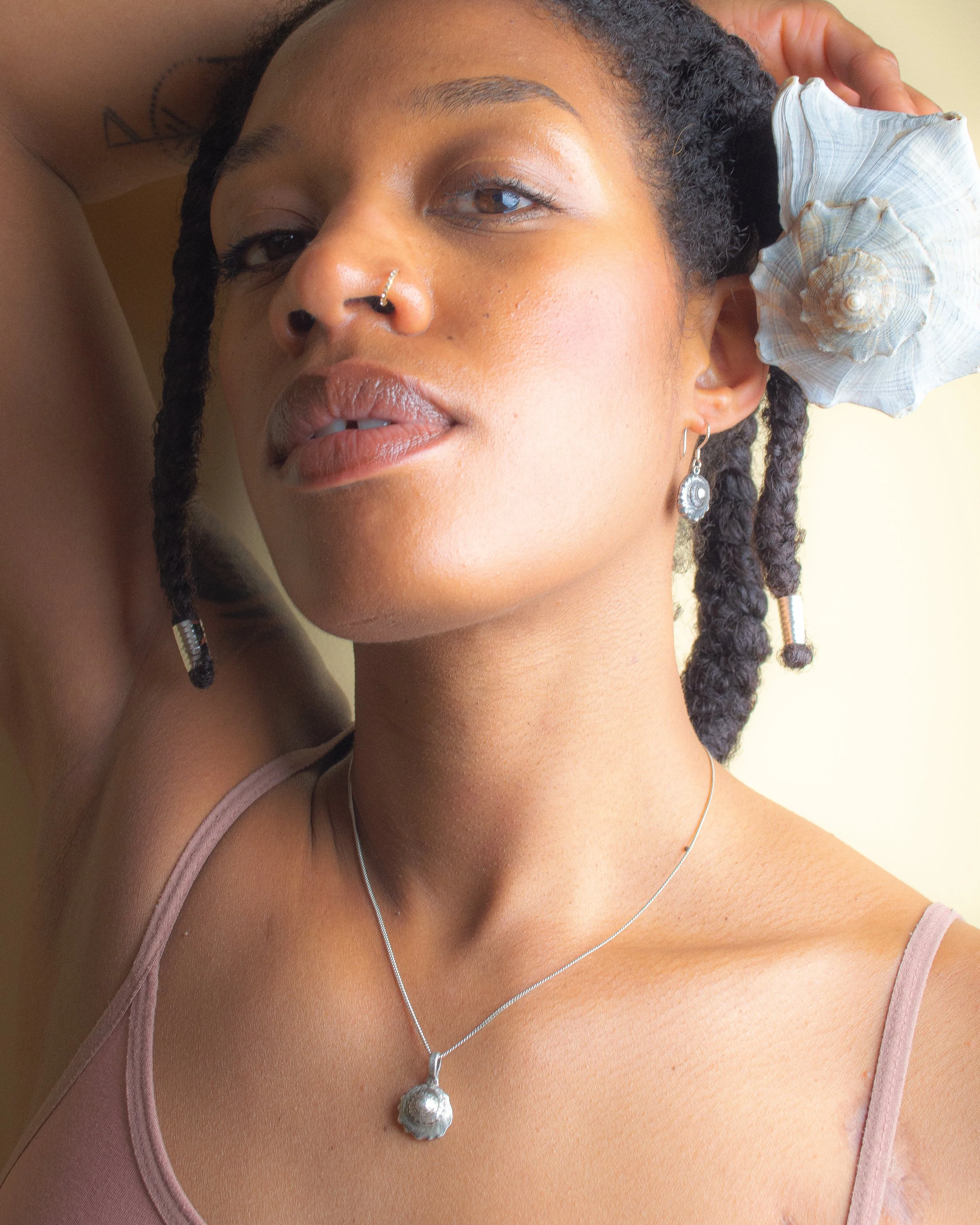 Fine silver round shell earring worn against bare skin, with a large grey scallop shell in the background