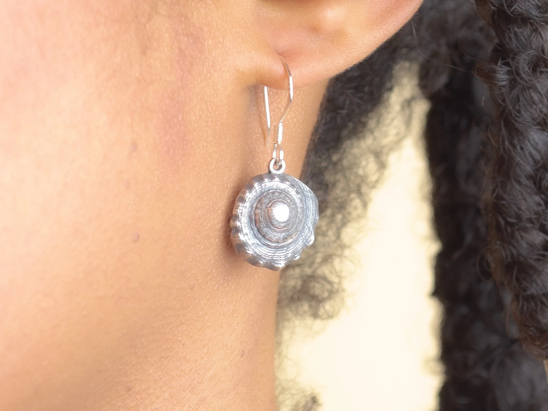 Fine silver round shell earring worn against bare skin, with a large grey scallop shell in the background