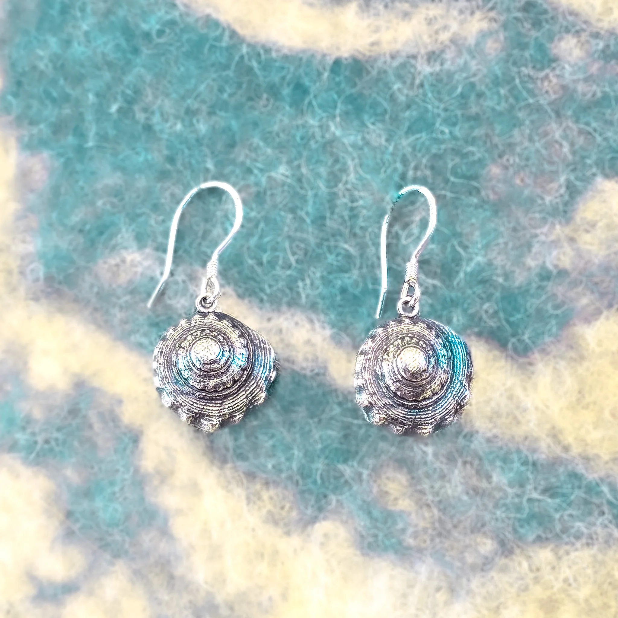 Fine silver round shell earring worn against bare skin, with a large grey scallop shell in the background