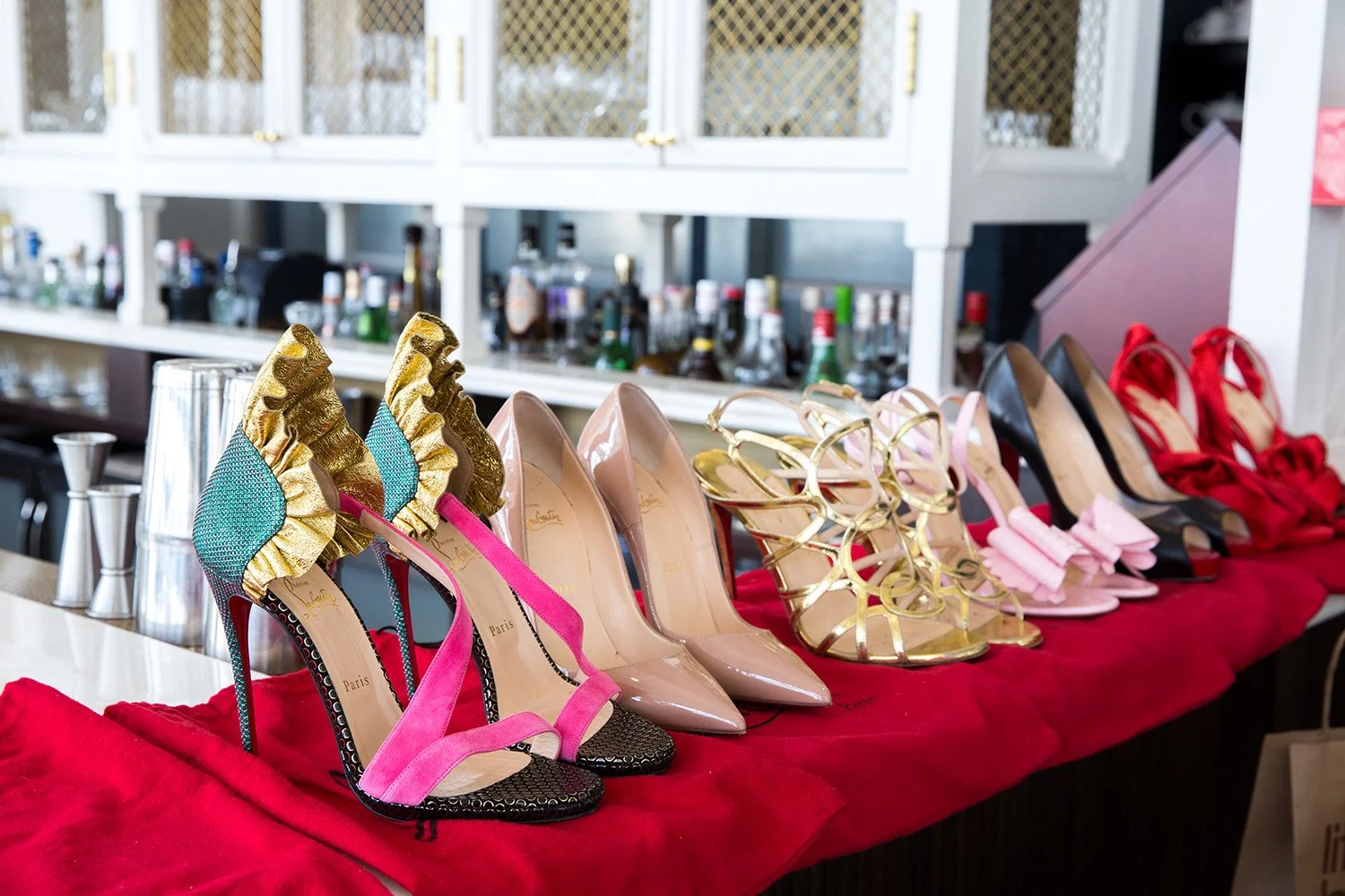A row of high-heeled shoes on a red cloth-covered surface, including designs with colorful ruffles, blush pink, black, gold with pink ribbons, pink bows, and red satin styles, with a bar in the background.
