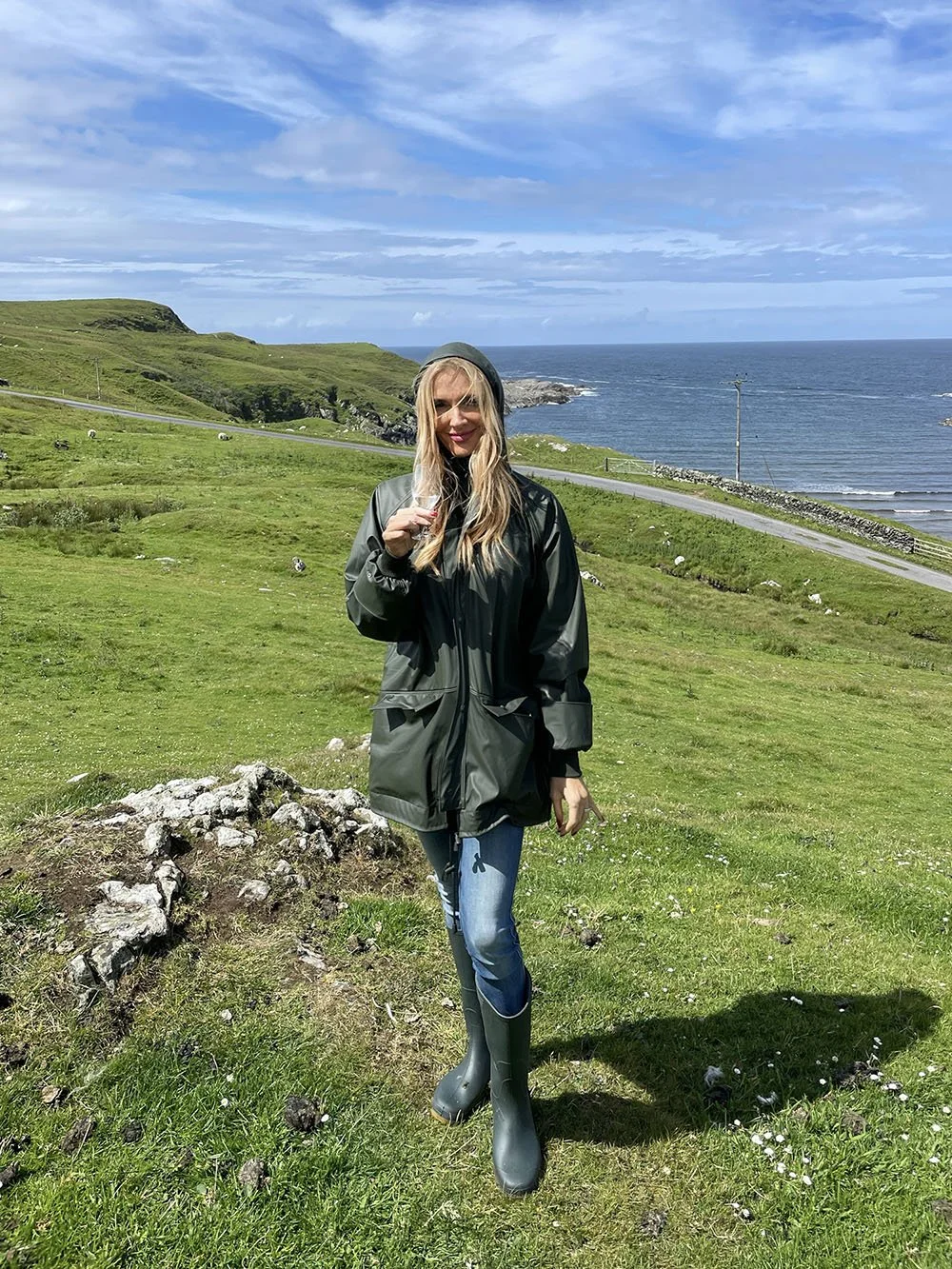 A woman in a green raincoat, jeans, and rain boots standing on a grassy field near the coast, holding a glass, with green hills and the ocean in the background on a partly cloudy day.