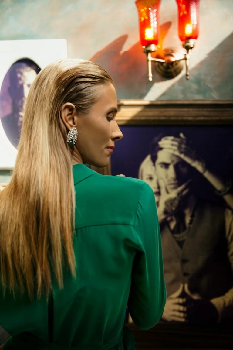 A woman with long blonde hair wearing a green top and large earrings is standing in front of a black and white portrait in an indoor setting, possibly a gallery or museum.