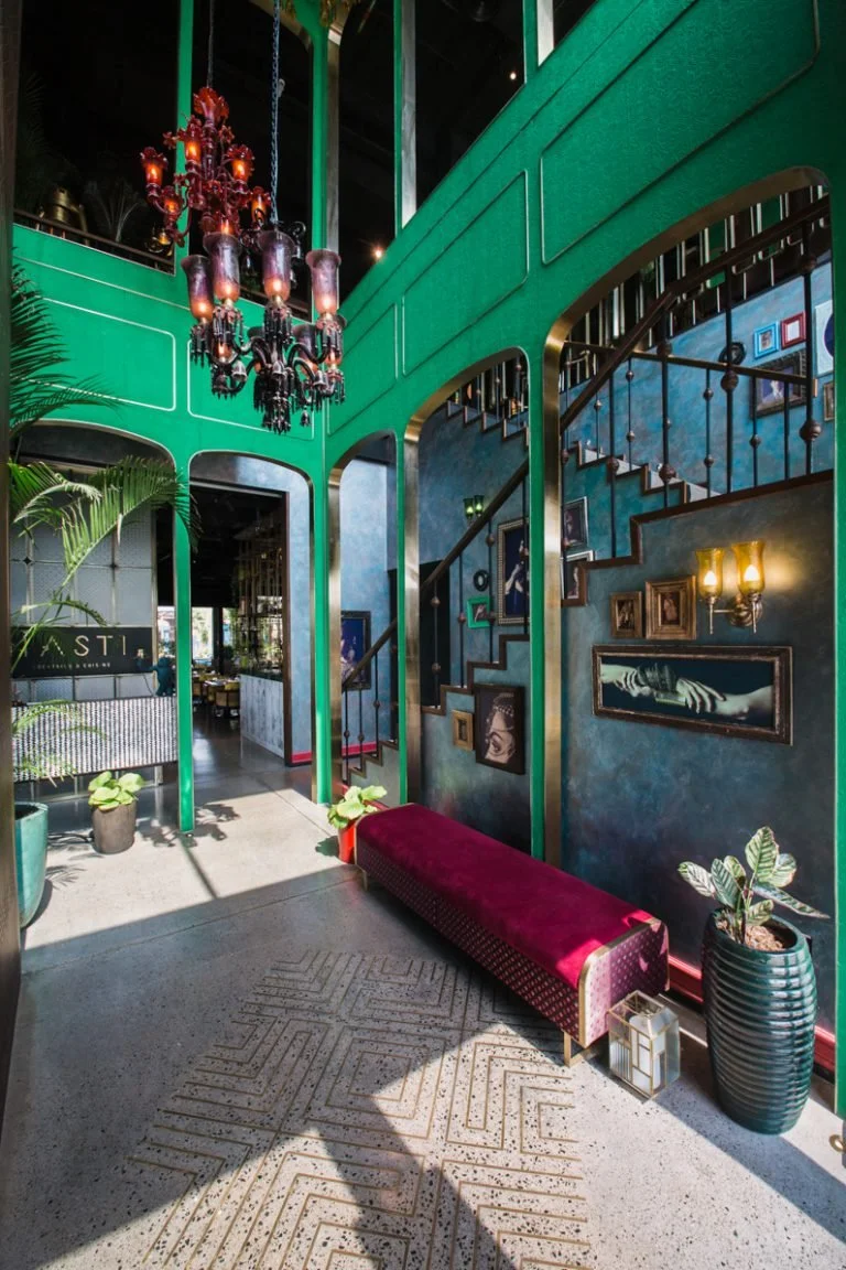 Interior of a stylish building with green walls, a red chandelier, framed artwork, a red velvet bench, potted plants, and a staircase.