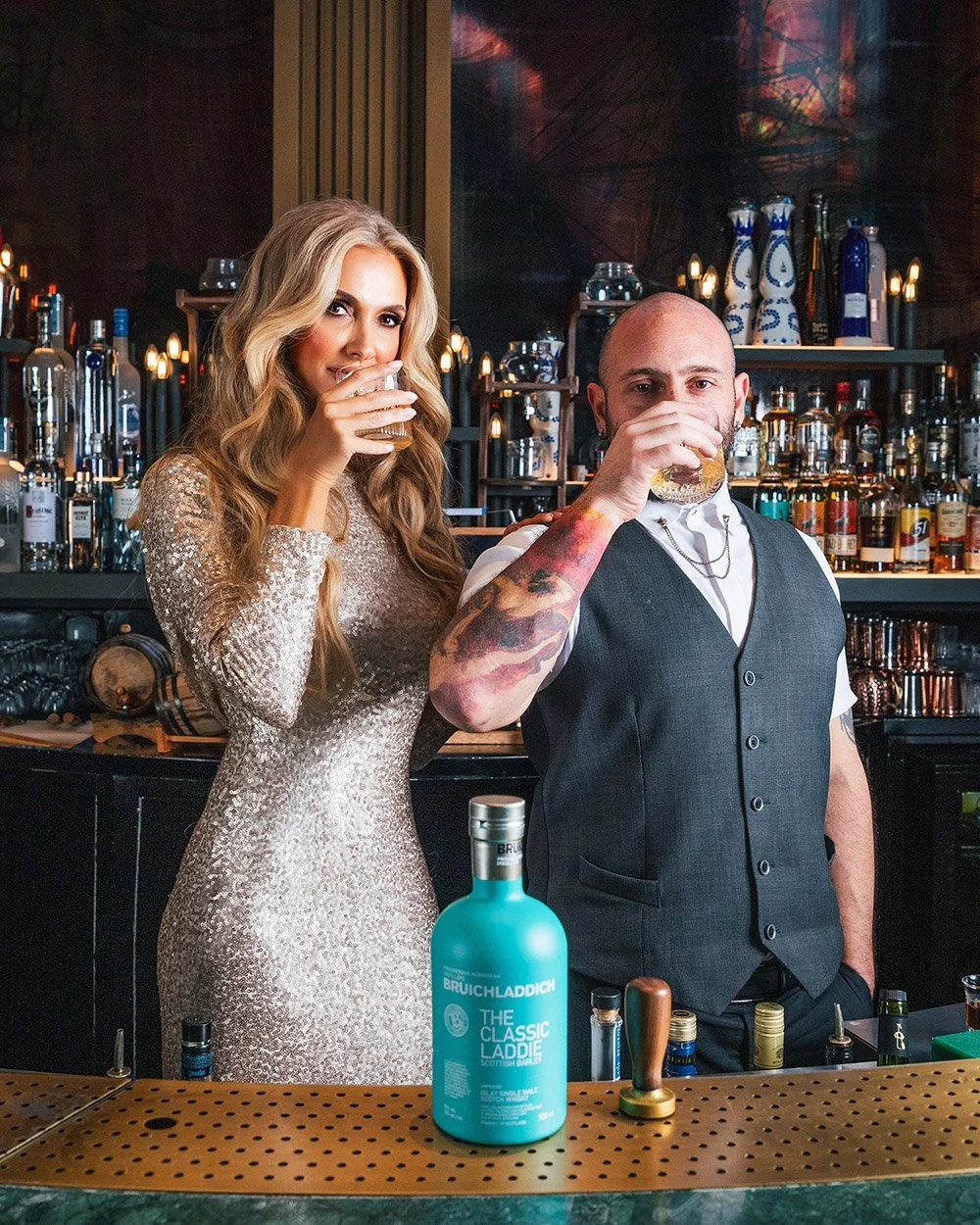 A woman in a shimmering dress and a man in a vest, both holding drinks, standing behind a bar with bottles of alcohol, glassware, and bar tools.